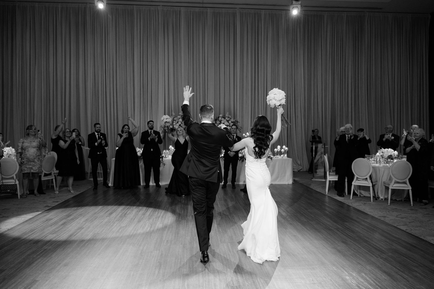 Black and white image of wedding couple making triumphant entrance with raised arms.