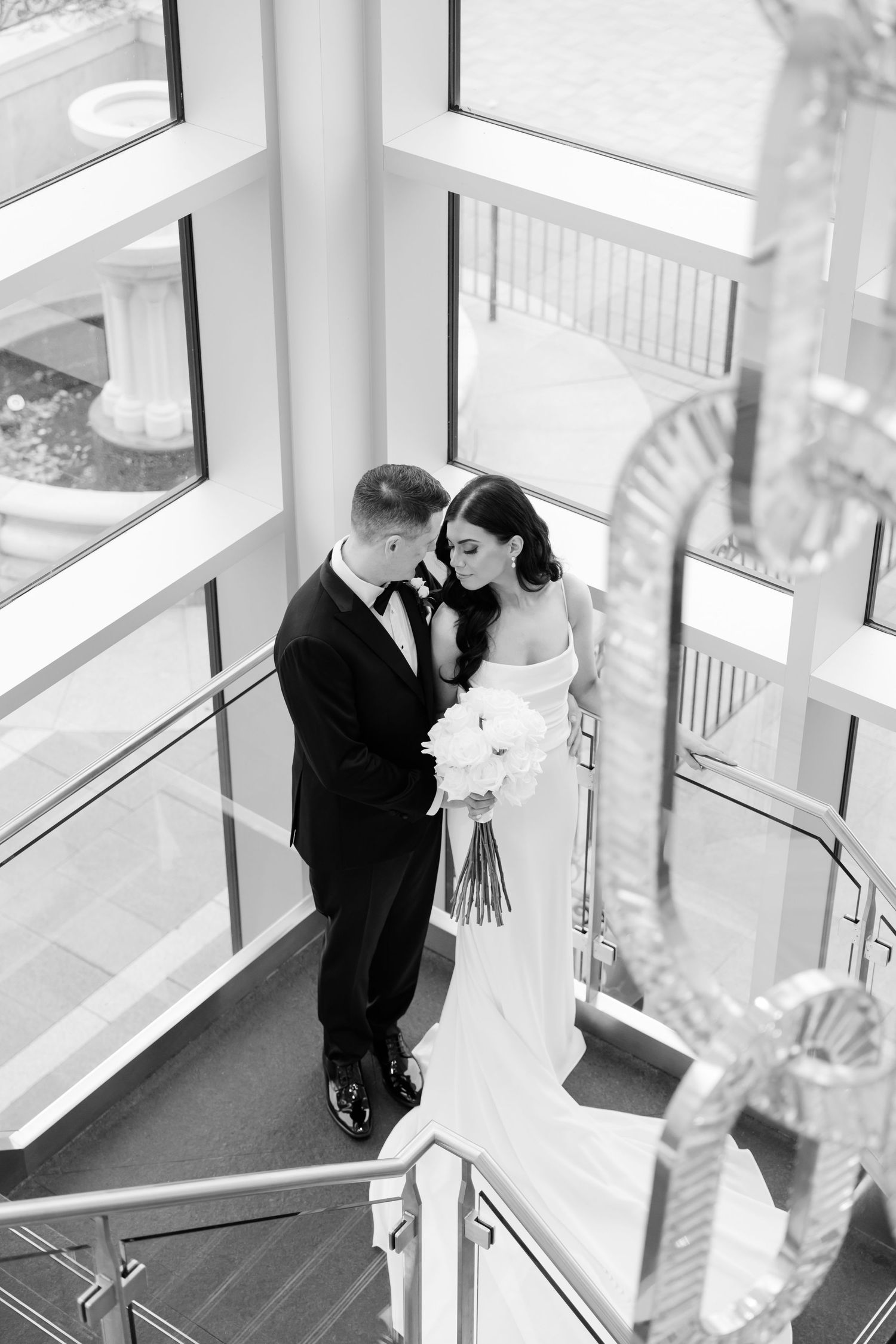Black and white photo of newlyweds sharing a tender moment on a staircase with large windows.