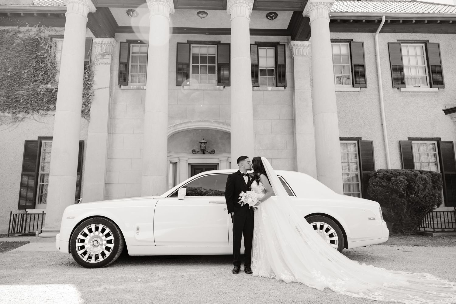 Black and white wedding photo featuring a couple with their luxury car against columned architecture.