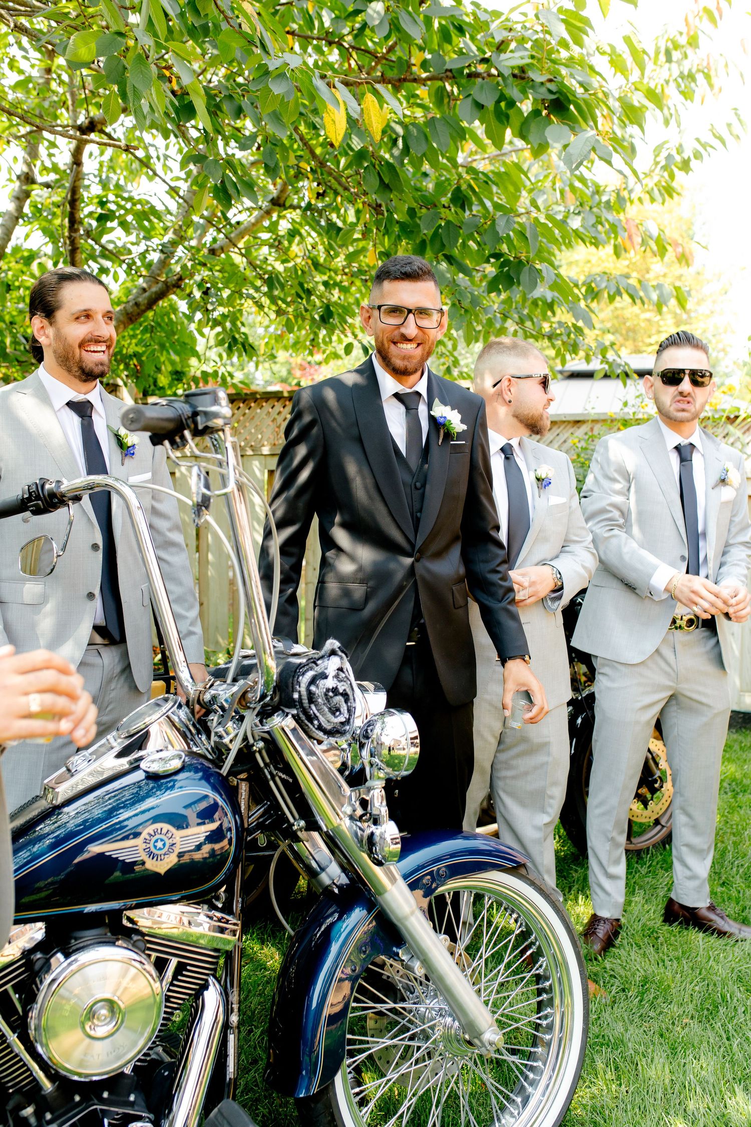 Groomsmen in light suits and sunglasses pose with classic motorcycles under trees.