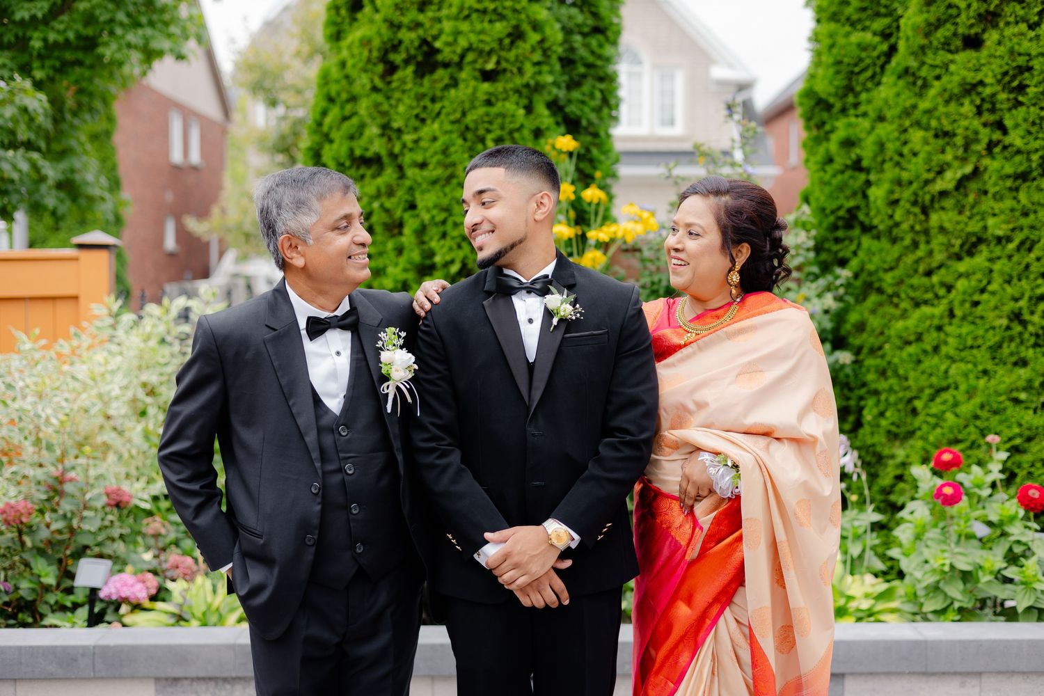 Wedding party members pose outdoors in formal attire against lush garden backdrop with vibrant flowers.