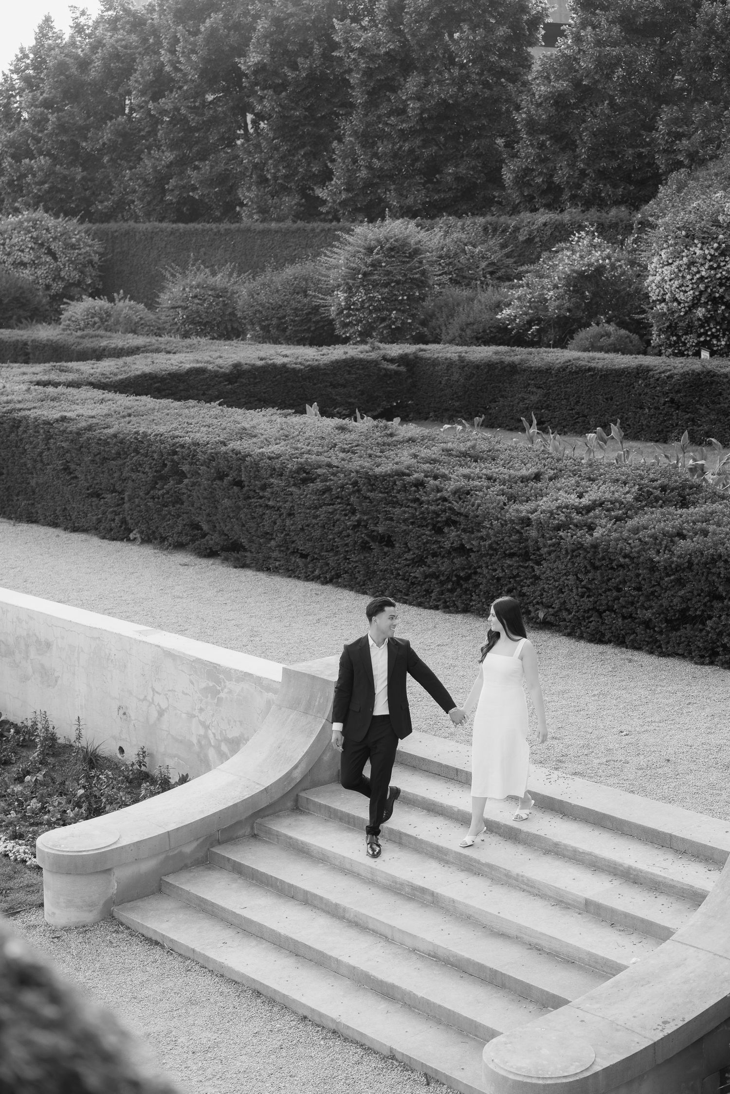 A black and white photo of a couple descending elegant curved stone steps in a formal garden.
