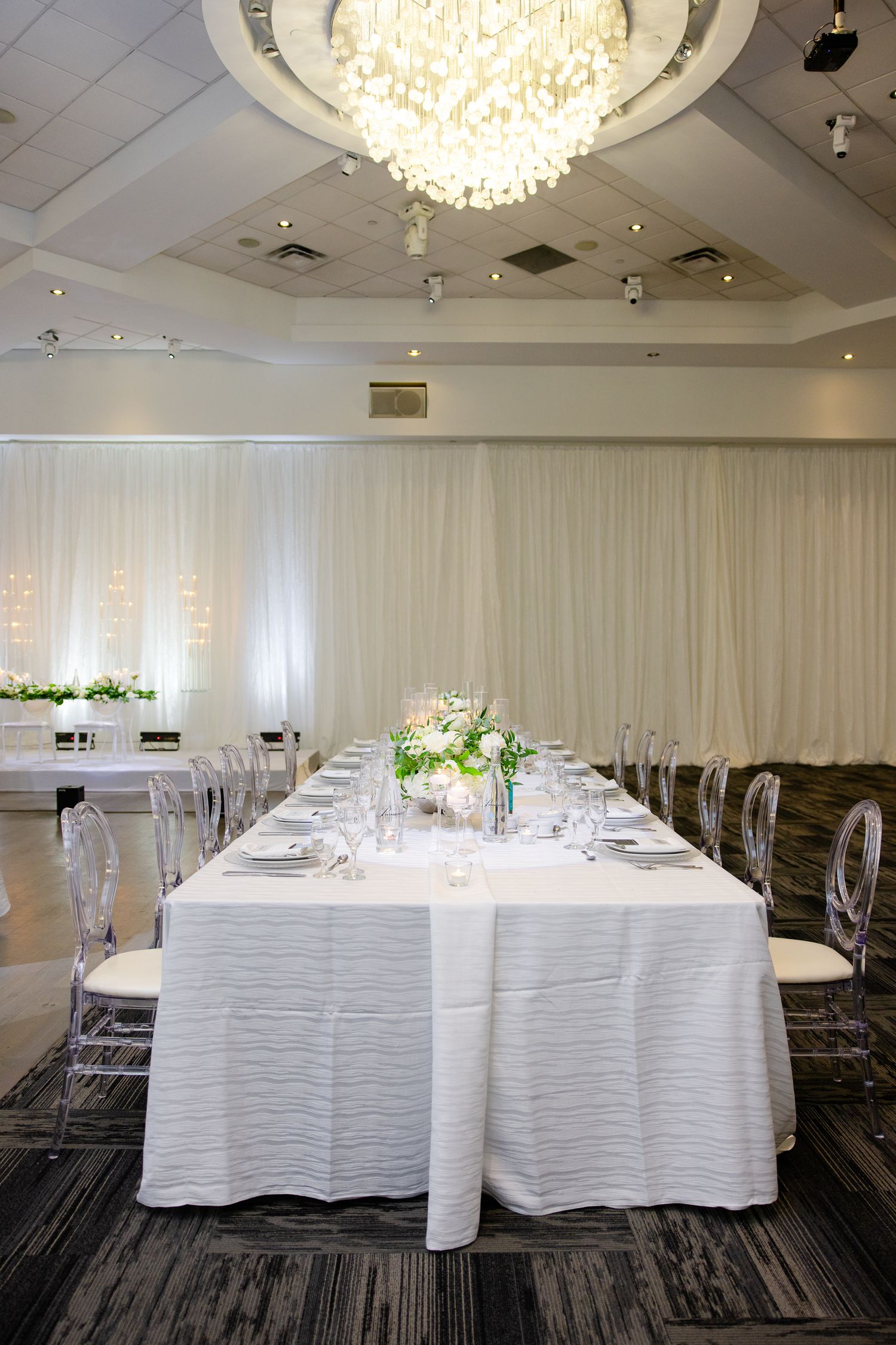 Elegant wedding reception table setup with white linens and crystal chandelier in modern venue.