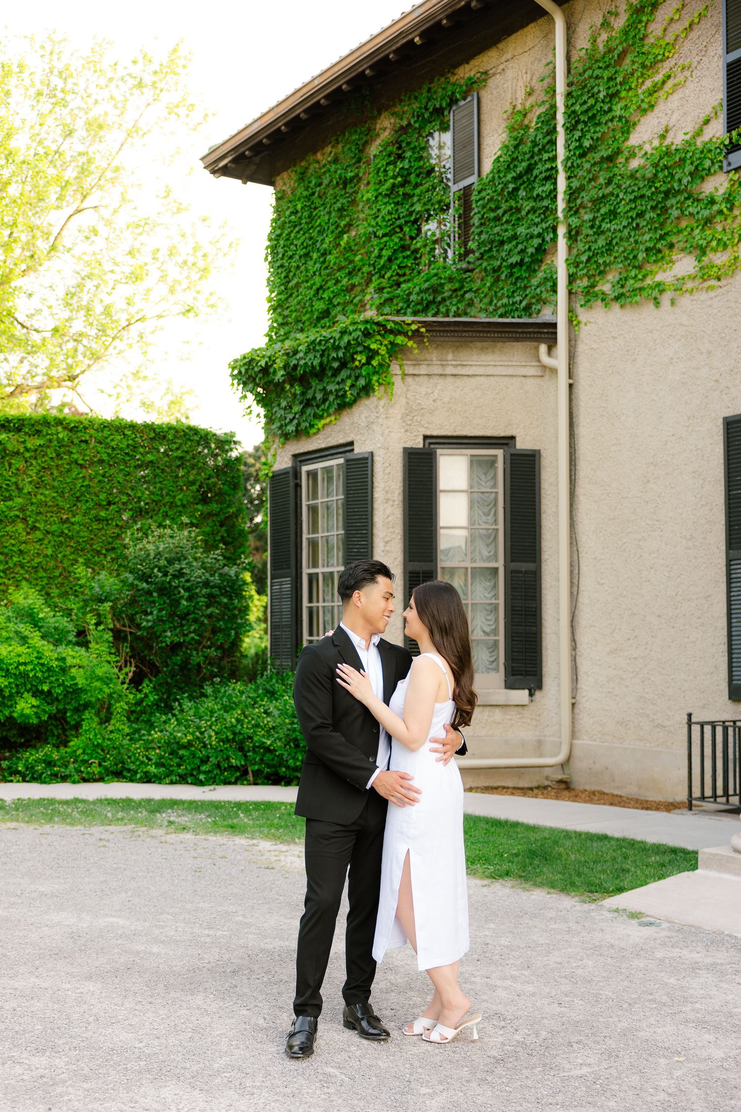 Romantic couple embracing outside ivy-covered mansion with black shuttered windows.