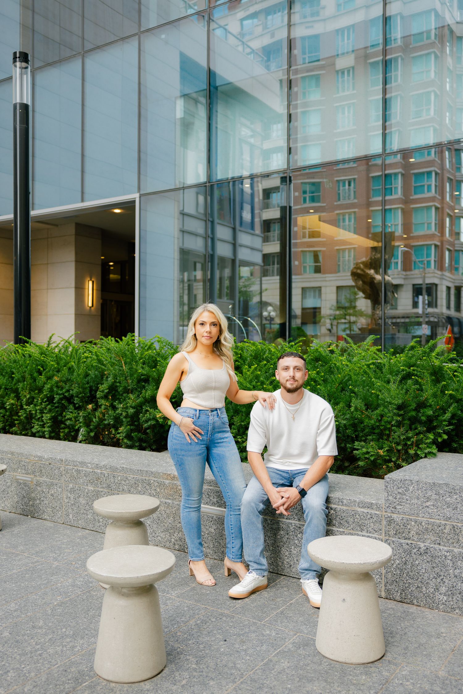 Stylish couple poses outside modern building with glass facade and decorative concrete bollards.