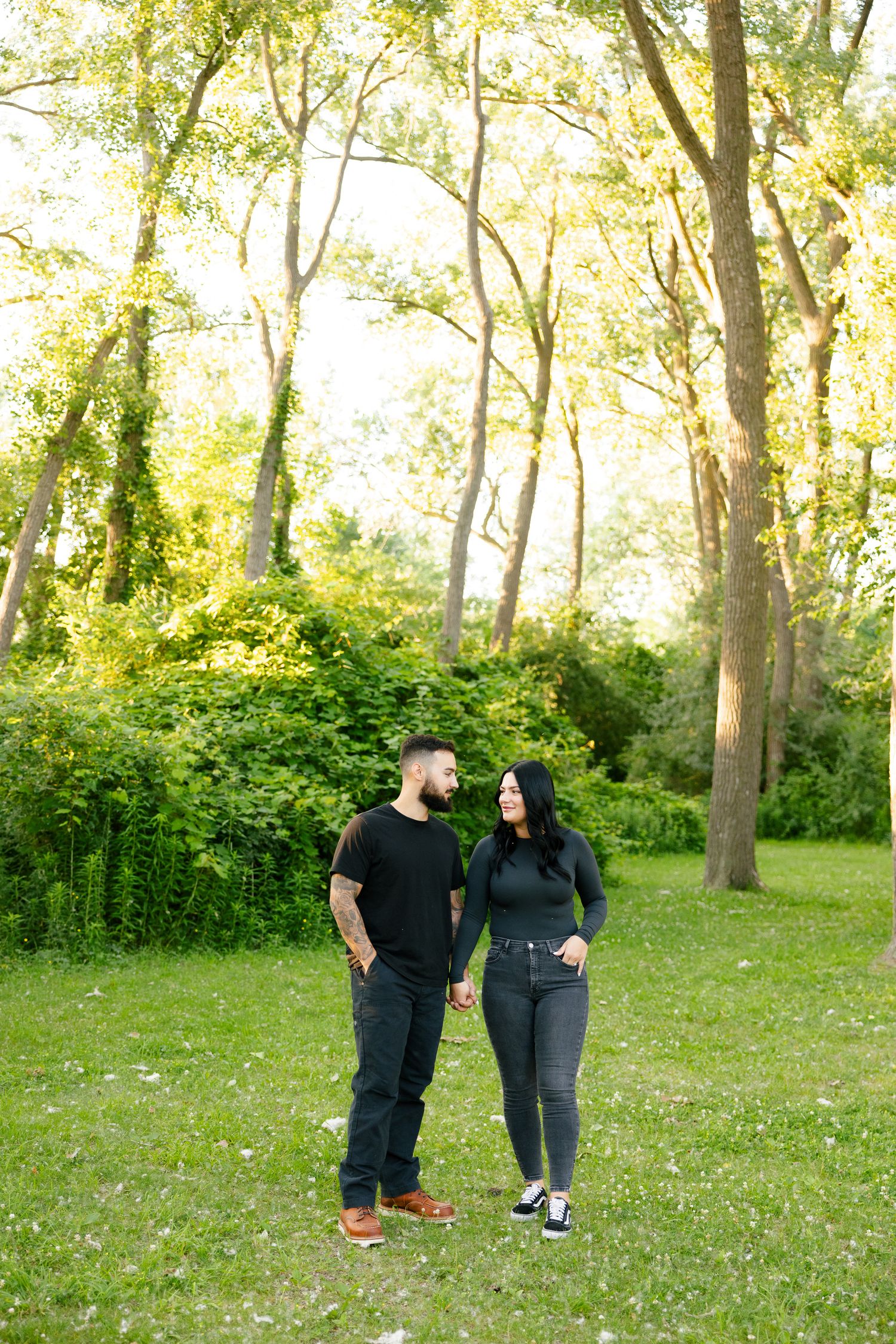 A couple stands together on a sunlit grassy area surrounded by tall trees.