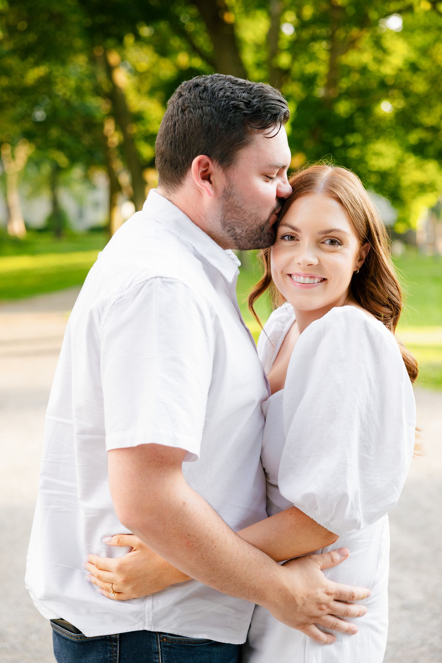 A loving couple embraces on a park path with sunlit trees in the background.