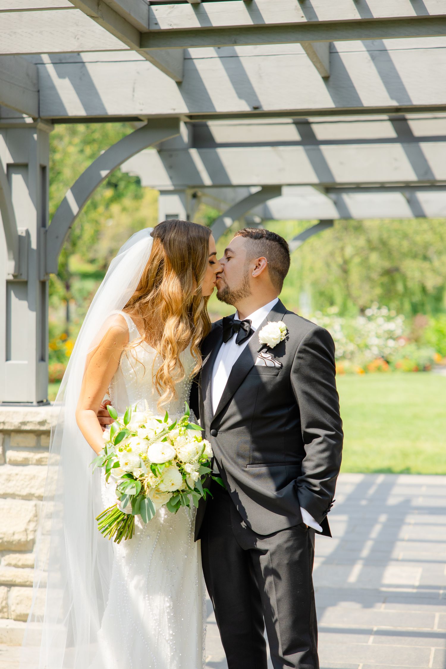 A romantic wedding moment under a white pergola with sunlit garden backdrop.