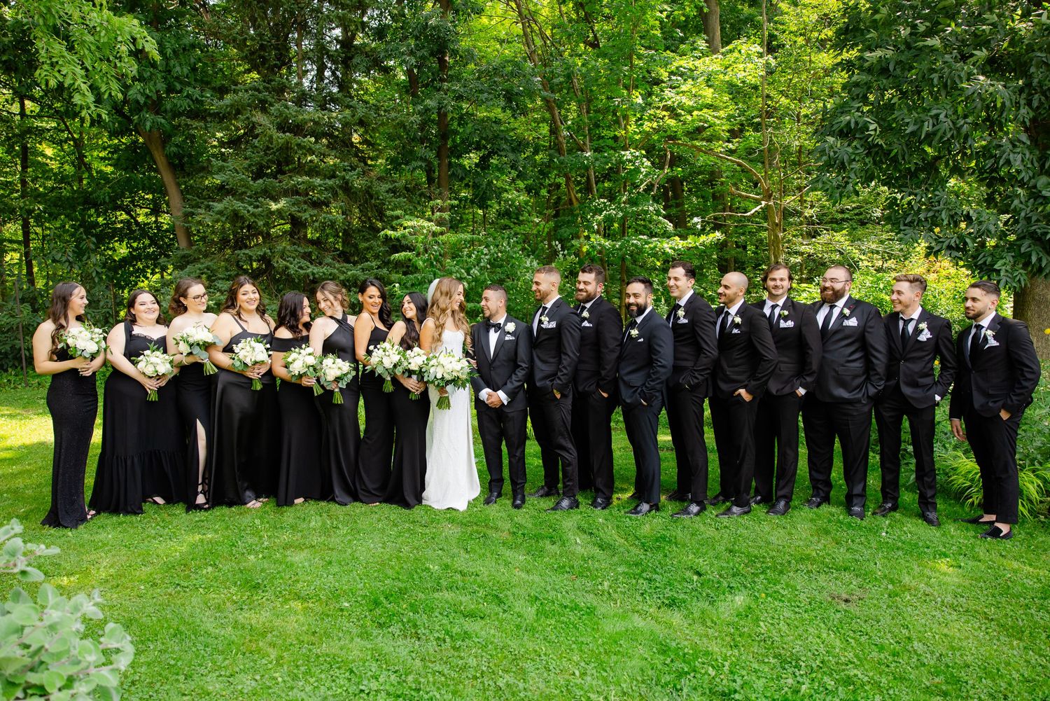 Wedding party in black dresses and tuxedos lines up for formal group photo on green lawn.