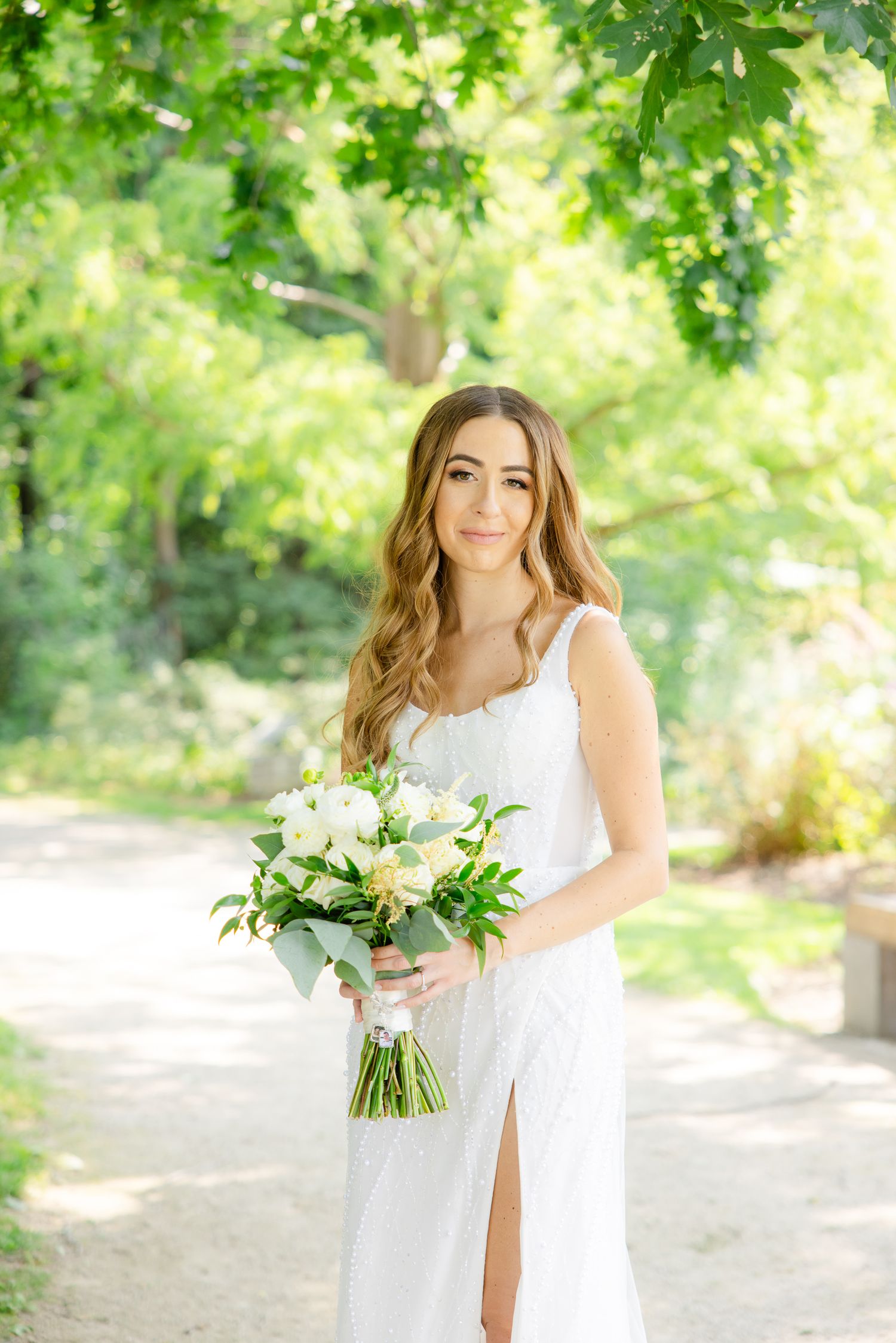 A bride in a white wedding dress holds a bouquet of white flowers in a sunlit garden setting.