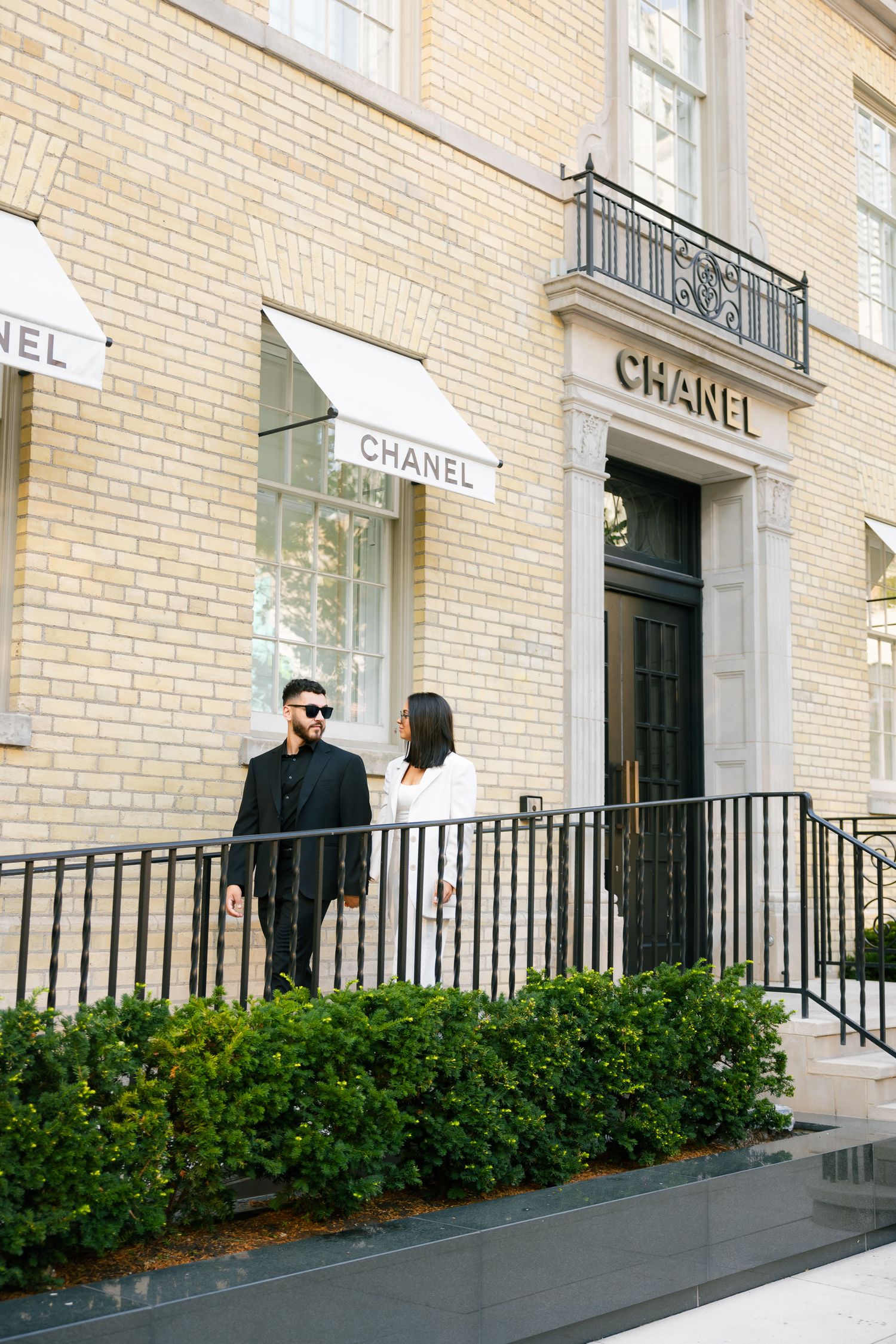 Elegant couple standing by Chanel storefront with white awnings and brick exterior.