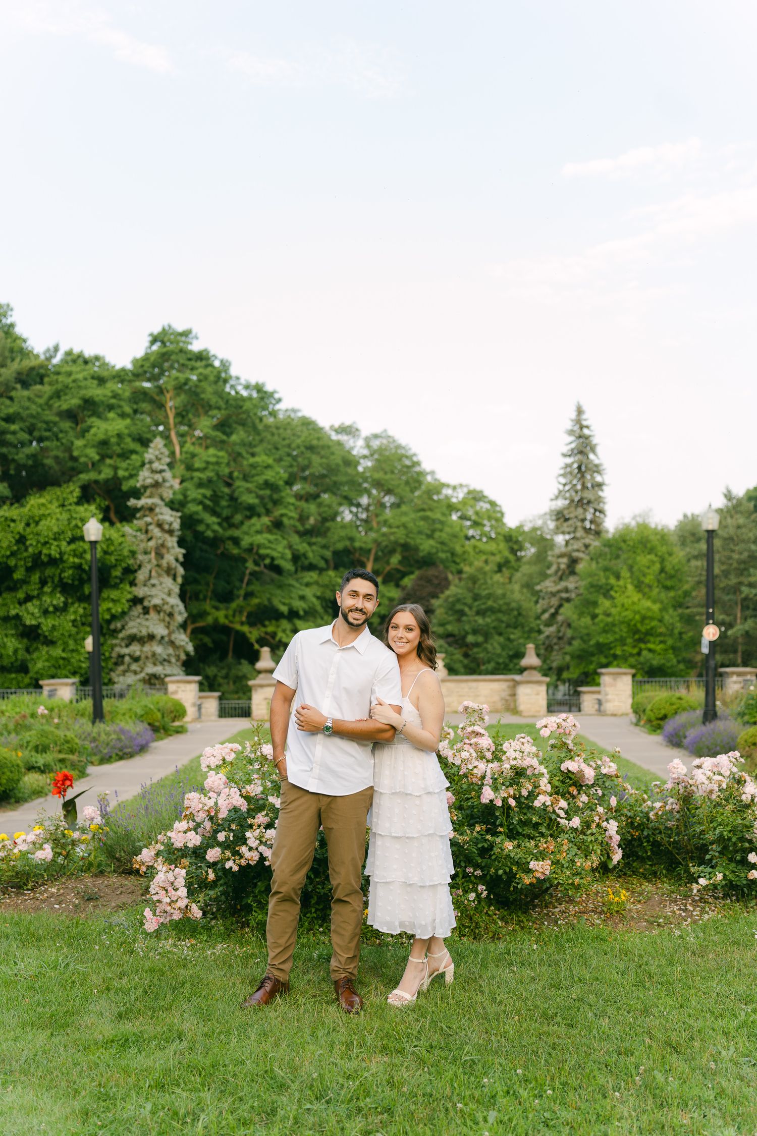 A full-length portrait of a couple standing in a manicured garden with pink roses.