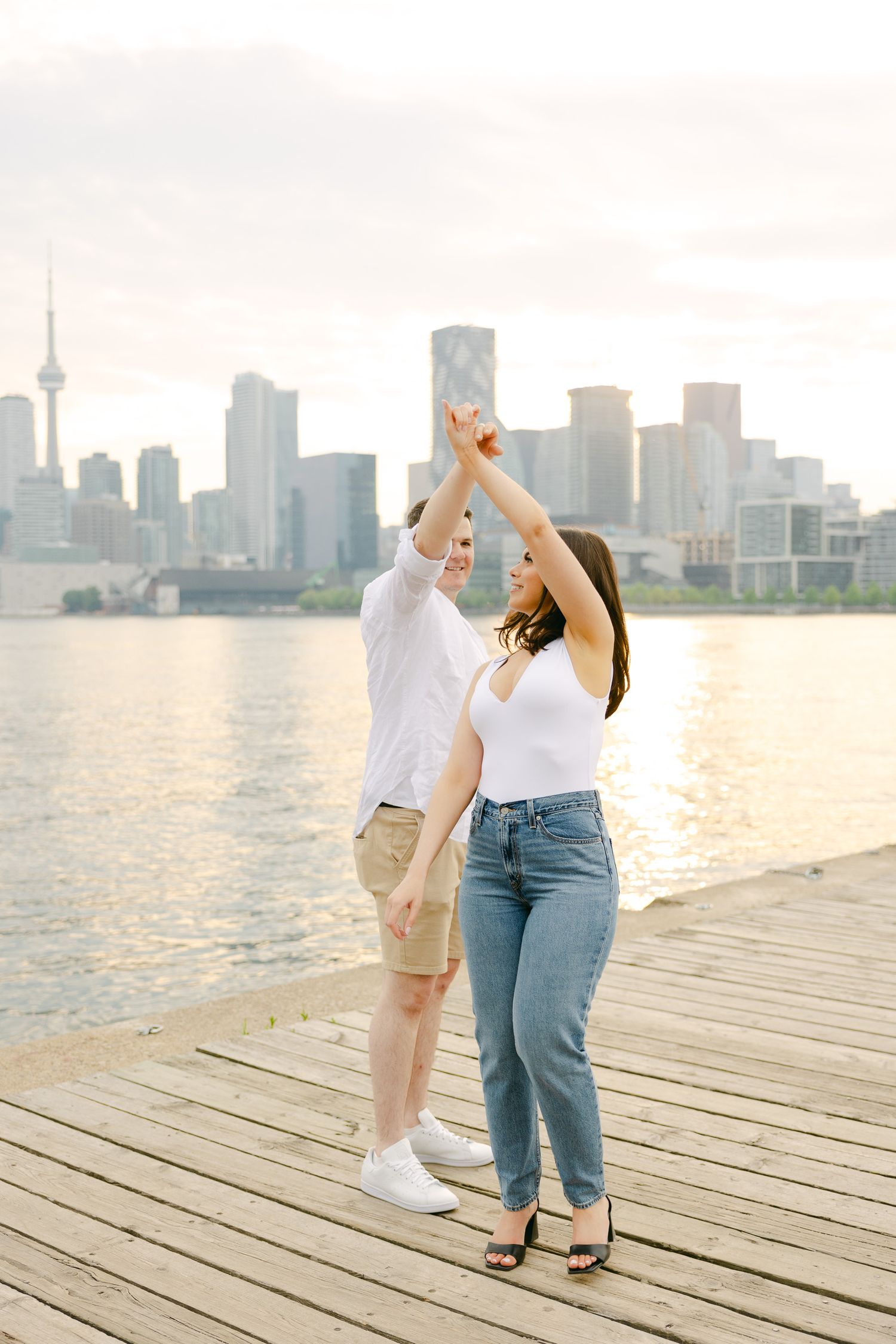 Dancing together on boardwalk with Toronto skyline silhouetted in soft light.