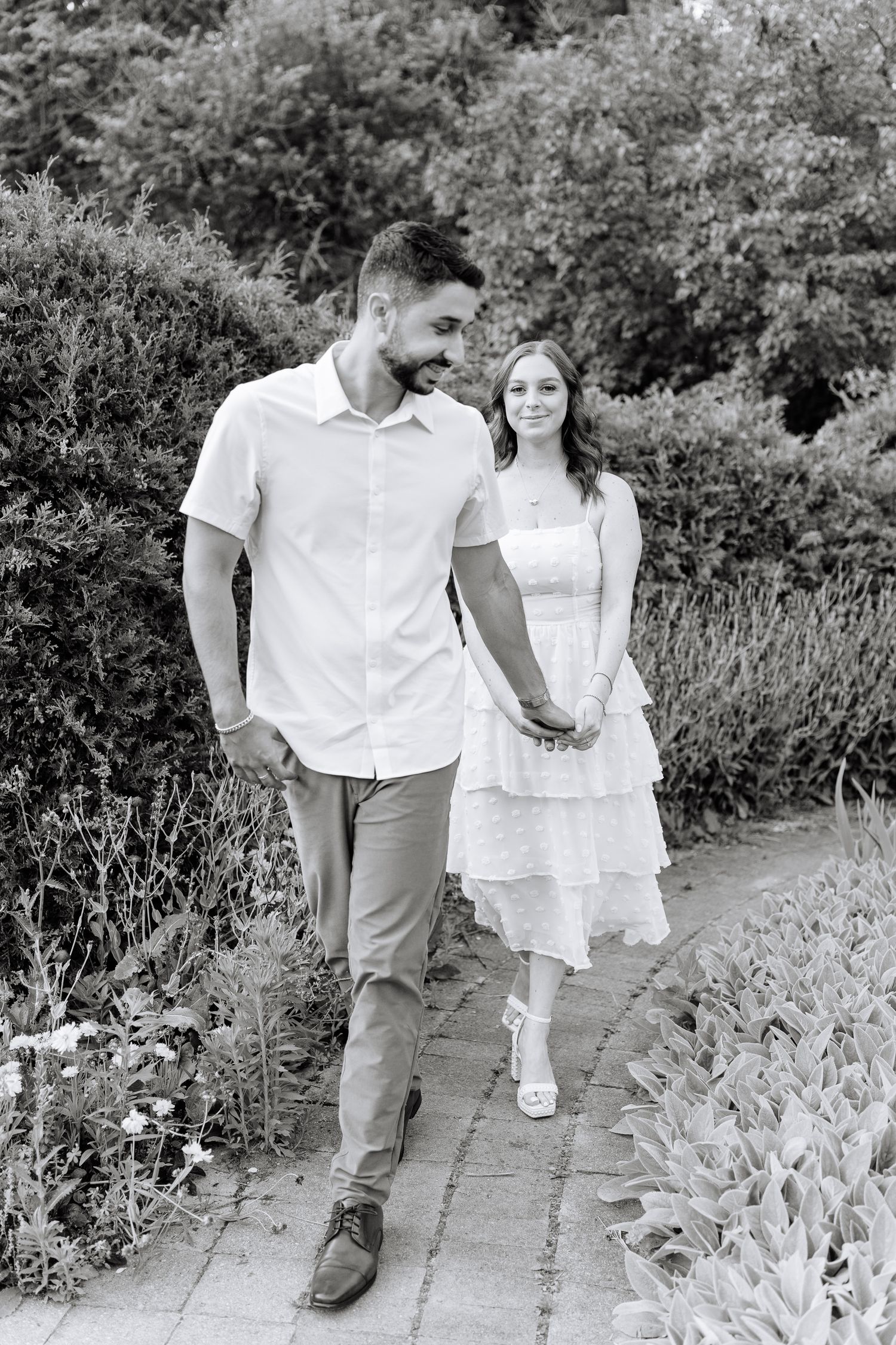 Black and white photo of couple strolling along garden pathway surrounded by plants.
