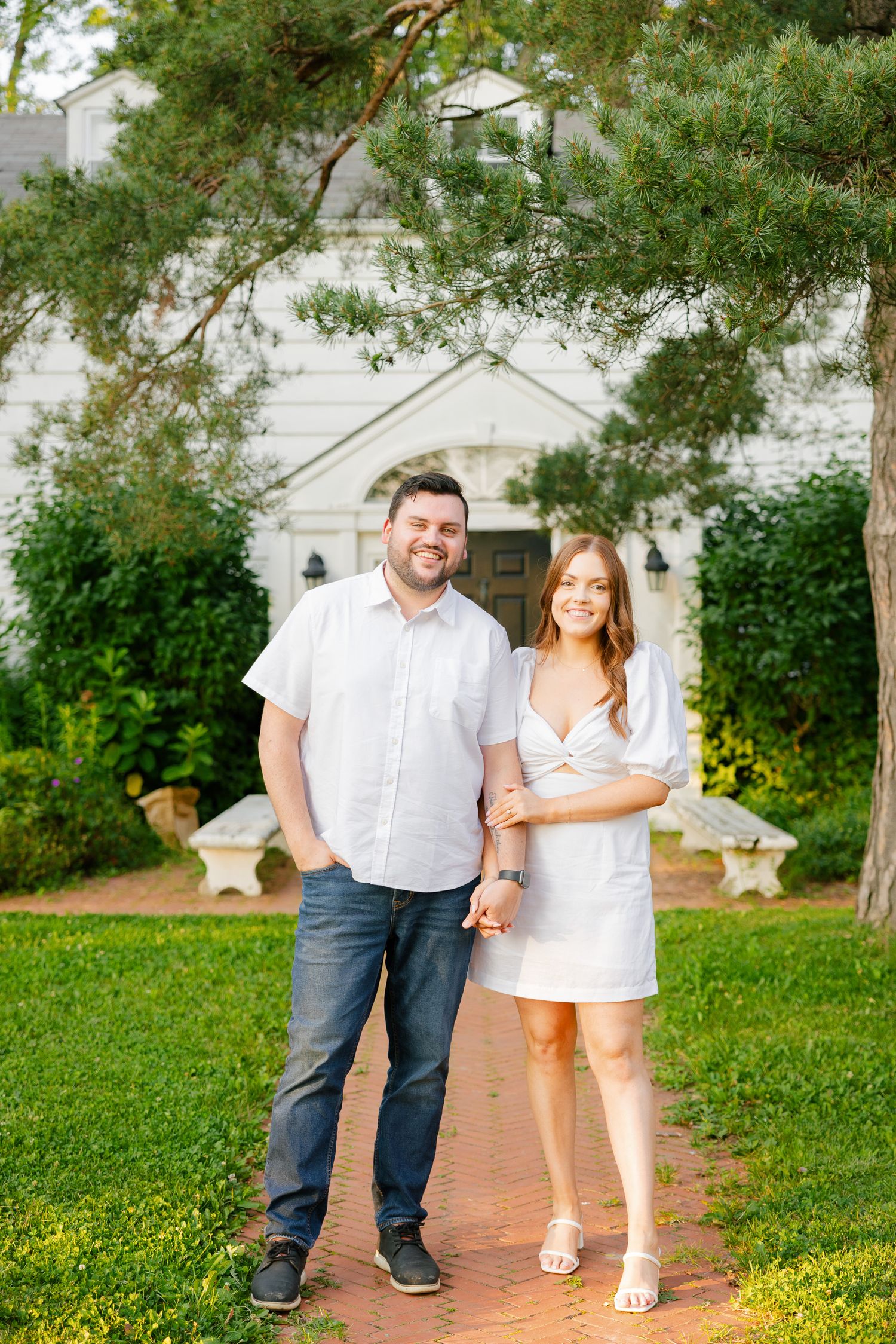 A couple stands together on a brick pathway in front of a white chapel with pine trees overhead.
