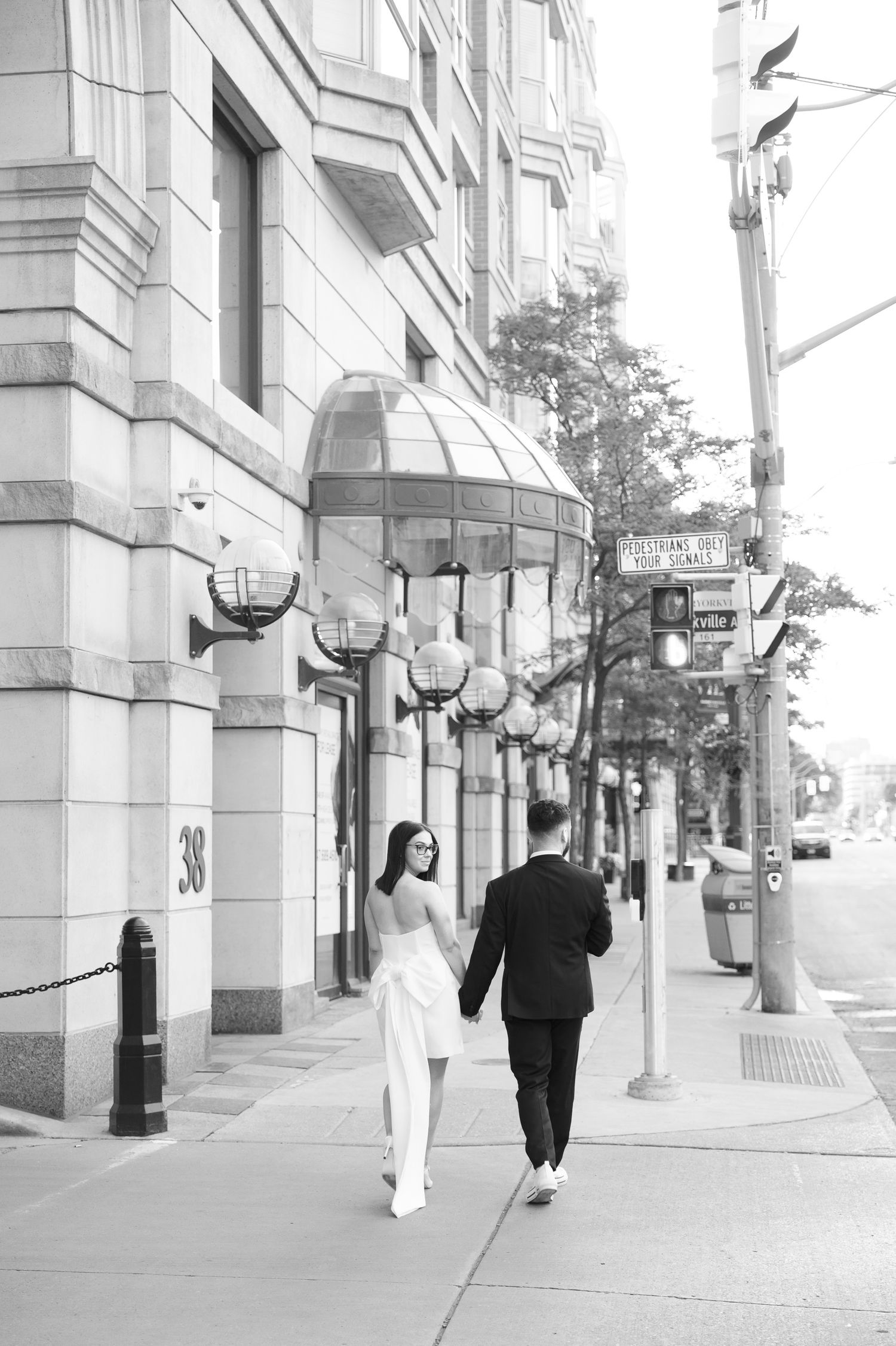 A couple in formal attire walks down a city sidewalk with classic street lamps and architectural details.