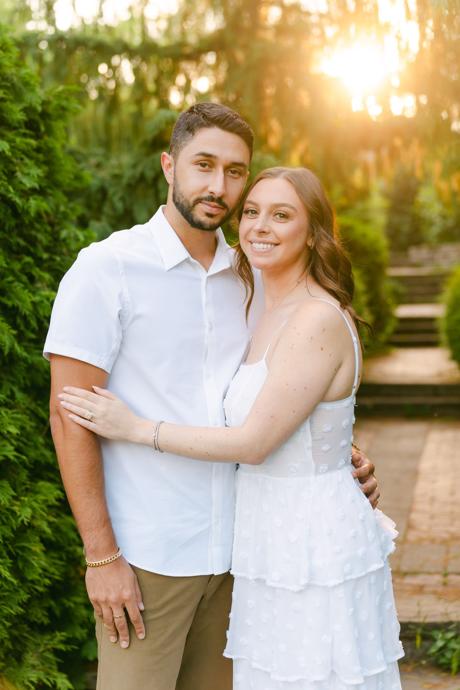 Couple in coordinating white outfits embrace on a garden path during golden hour.