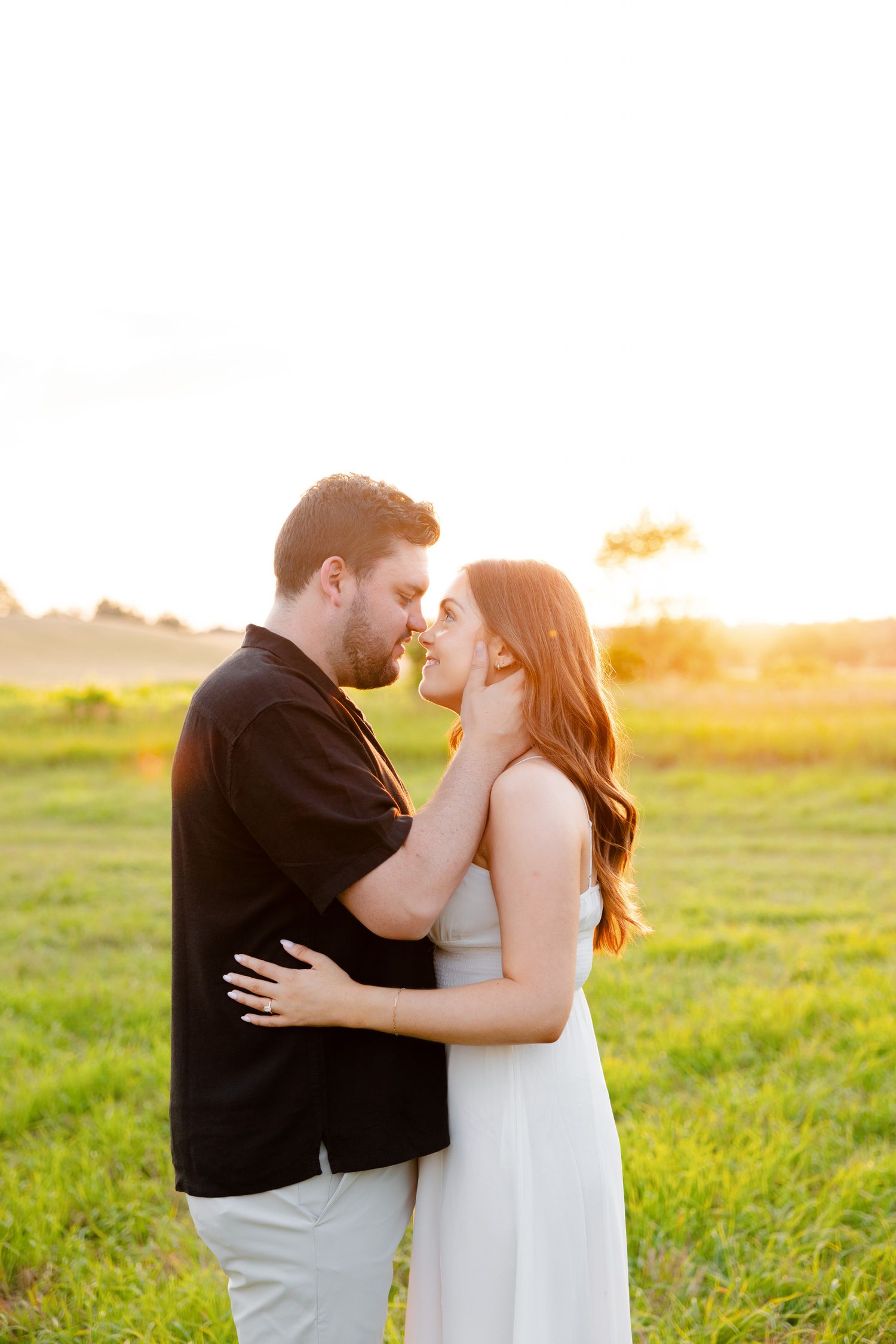 Couple shares an intimate moment during golden hour engagement photoshoot in a grassy field.
