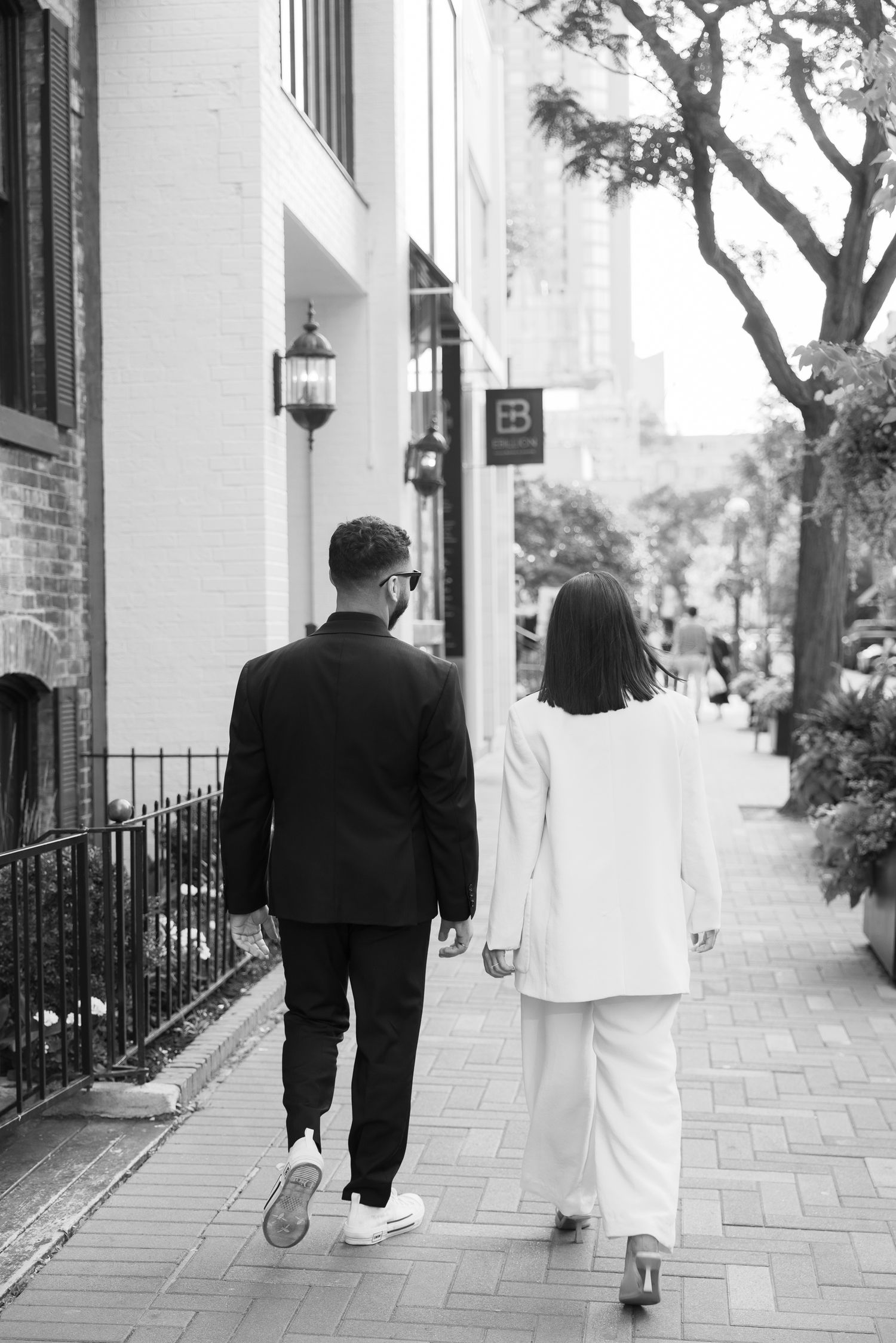 A black and white image of a couple walking down a city sidewalk with lanterns on the wall.