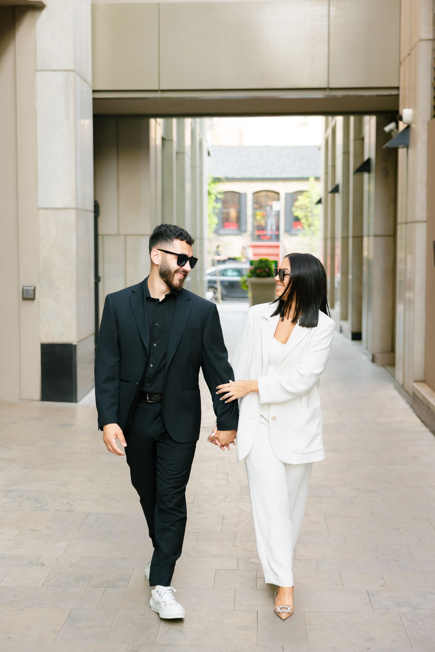 A couple in contrasting black and white outfits walks through a modern urban corridor.