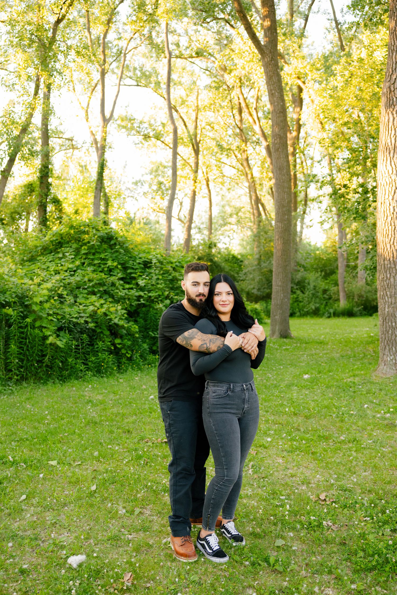 Full length portrait of couple embracing in sunny woodland clearing with tall trees.