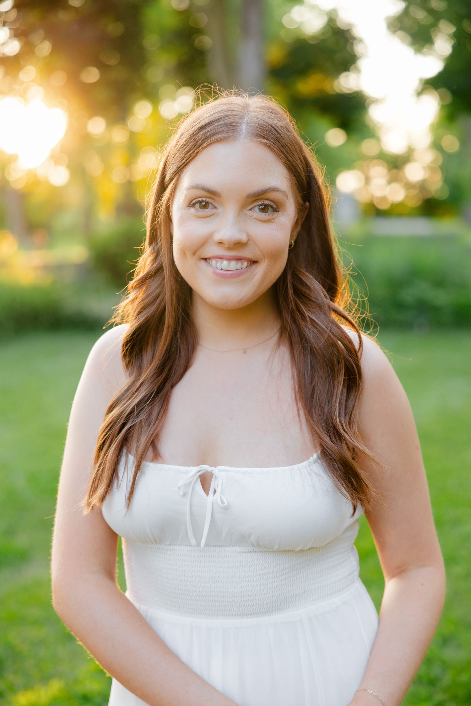 A woman in a white summer dress smiles during sunset in a garden setting.