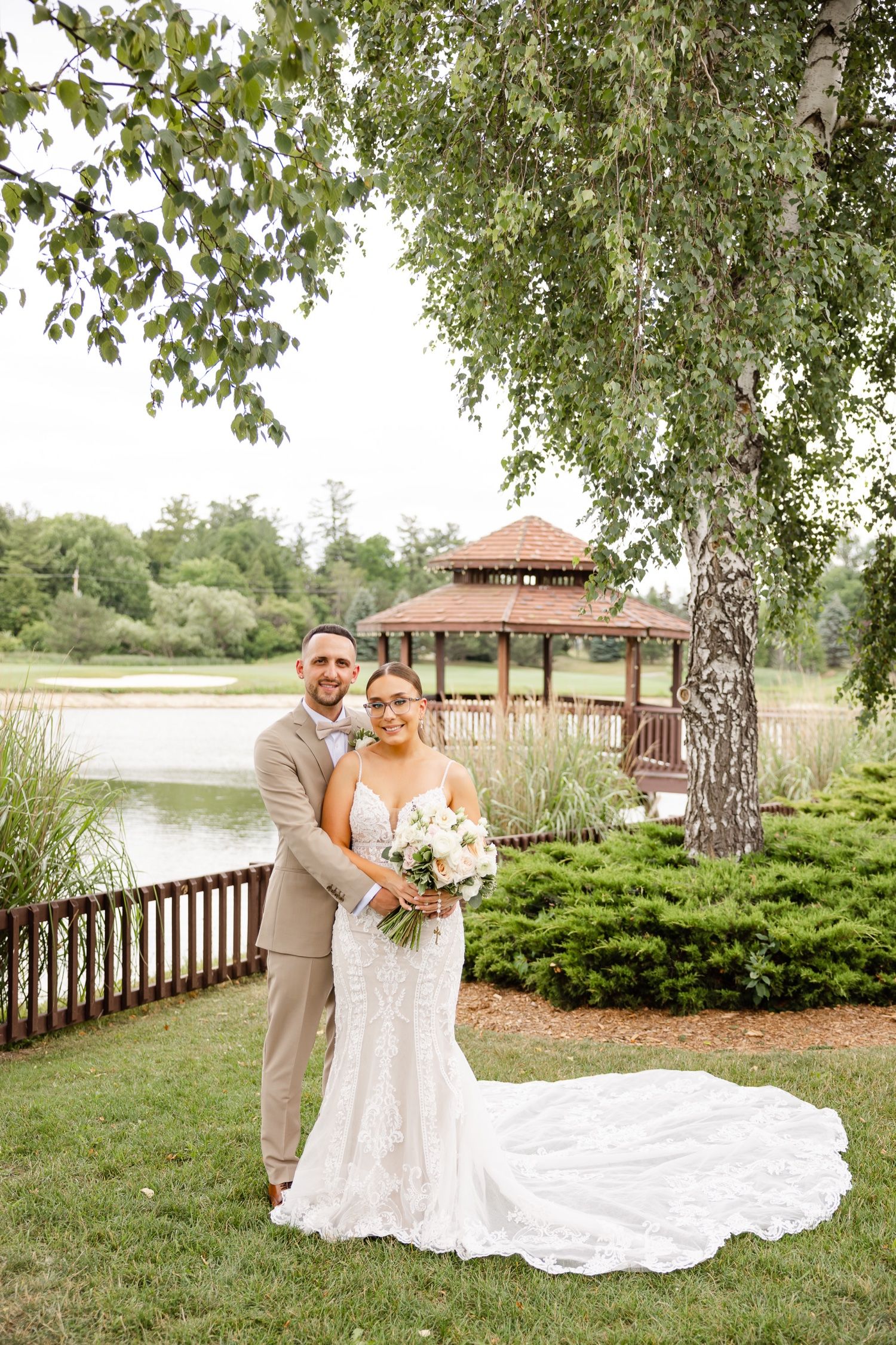 A wedding couple poses near a lake with a gazebo and birch trees in the background on their special day.