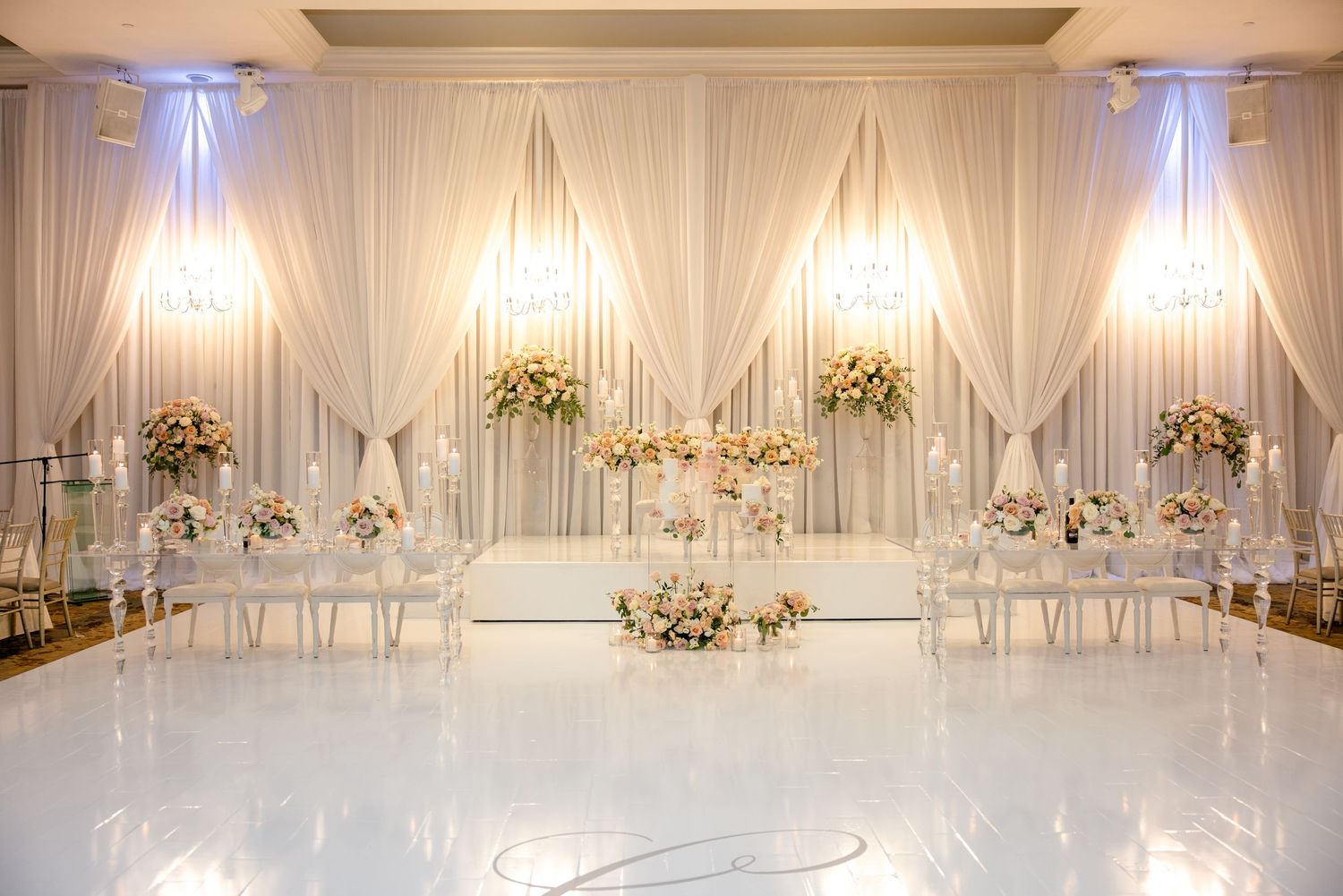 Grand ballroom wedding setup with white draping, floral arrangements and crystal chandeliers.