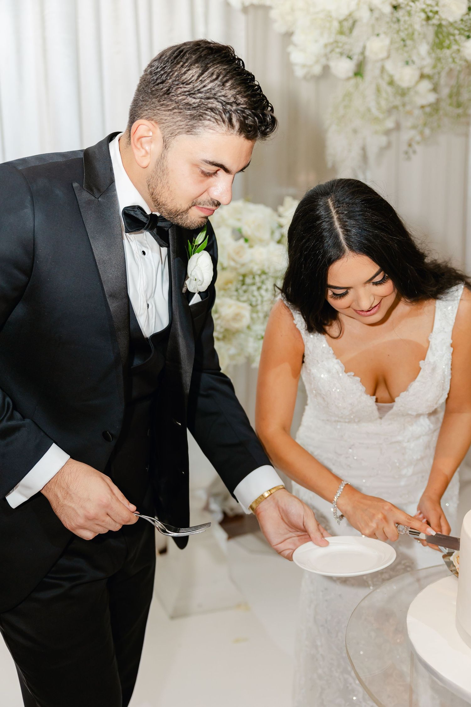 Wedding couple serving cake at their reception with white floral decorations.