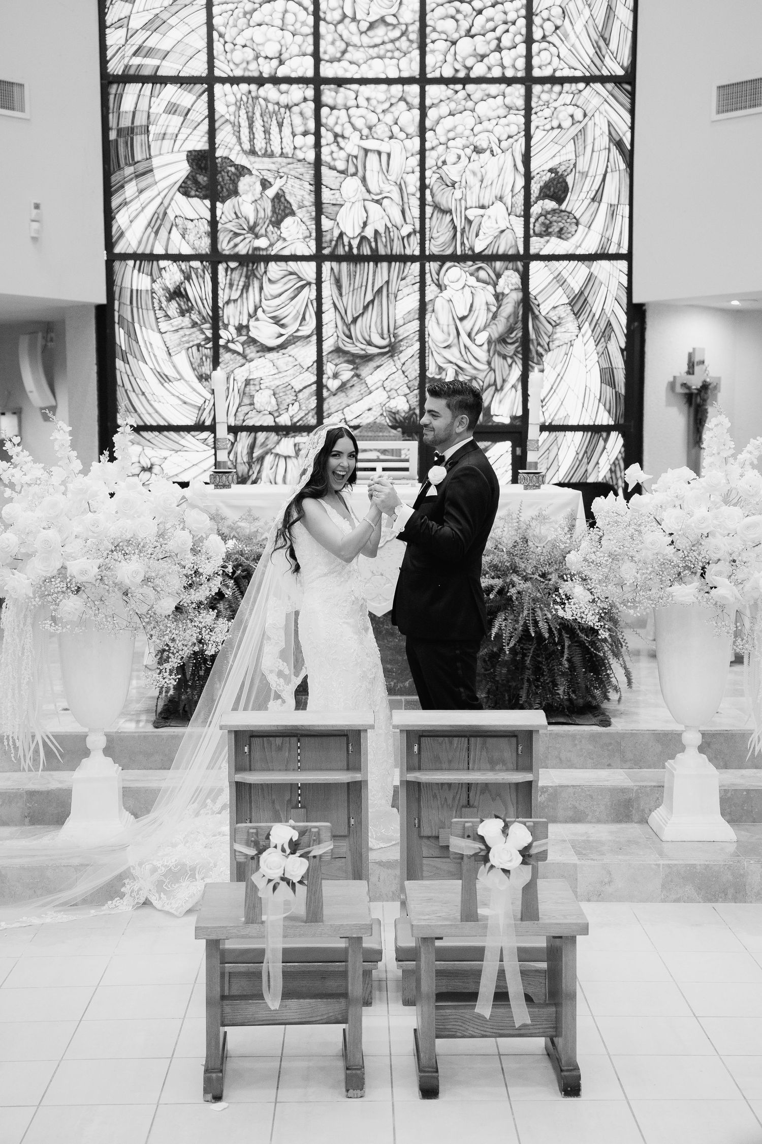 Black and white wedding photo with couple standing before artistic stained glass window and wooden chairs.