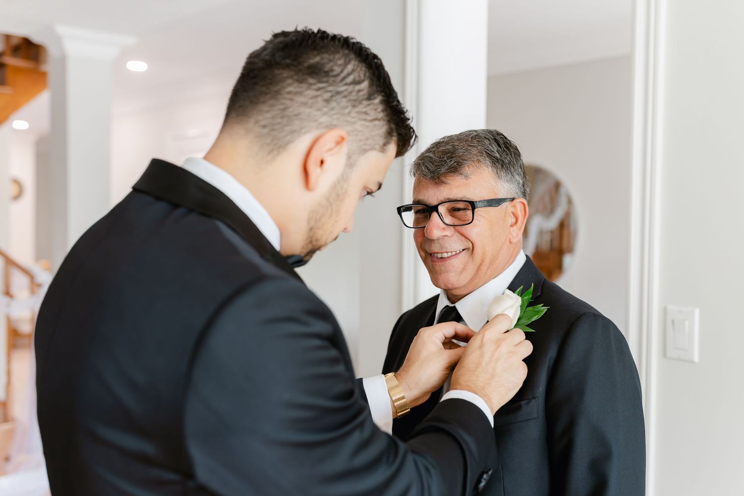 Two people in formal black attire share a moment while adjusting a boutonniere.