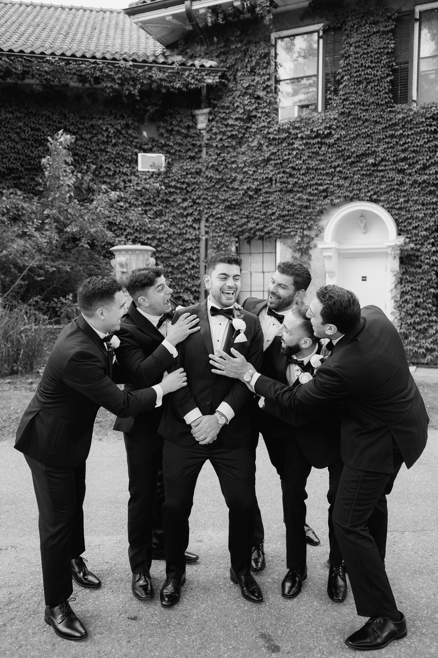 Black and white photo of groomsmen playfully gathered around in classic tuxedos.