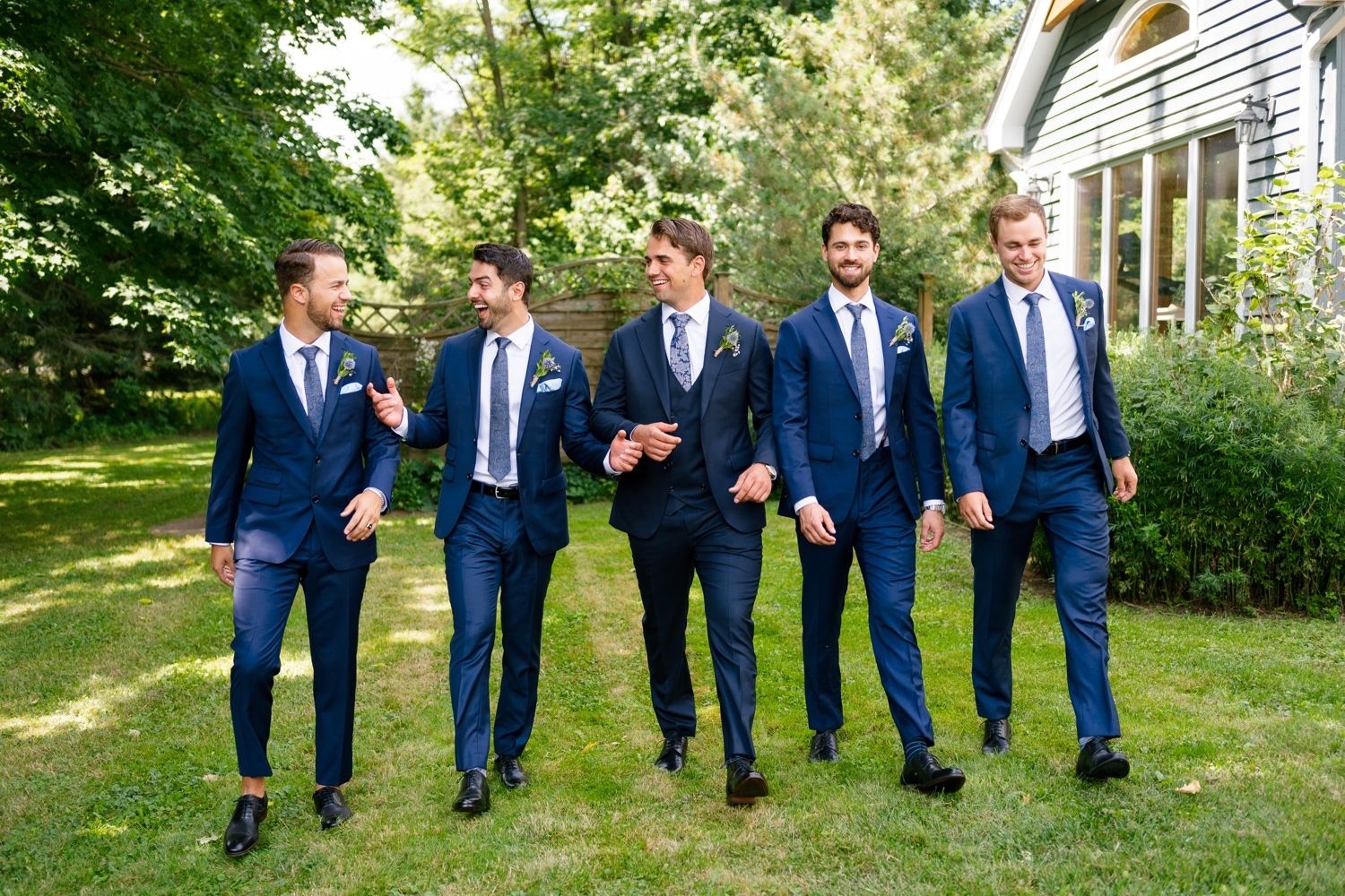 Wedding party in blue suits walking and chatting casually on garden path.