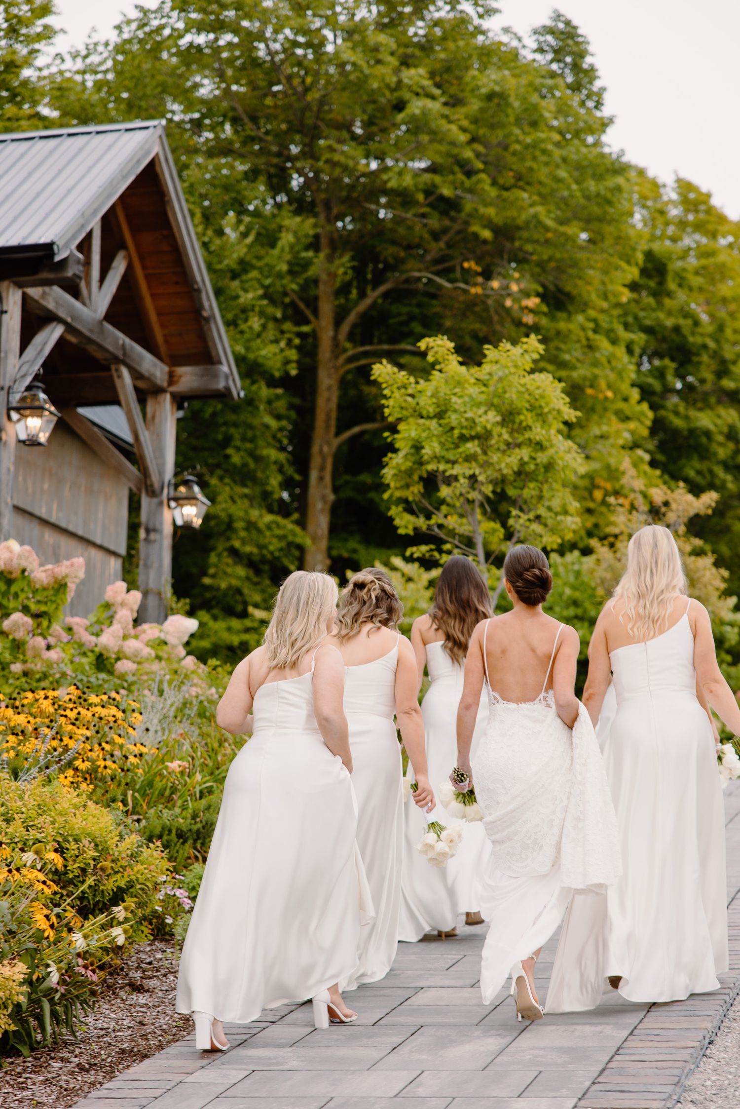 Bridesmaids in white dresses walking along a garden path near a rustic wooden structure.