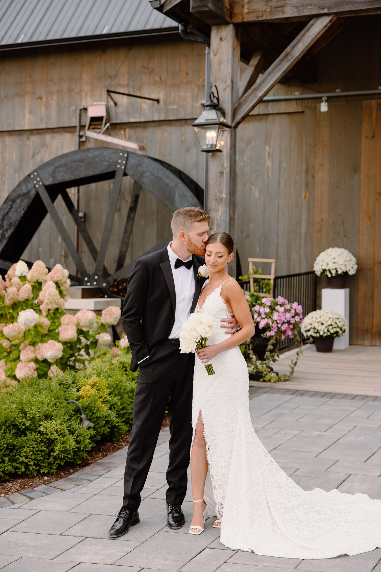 Elegant couple shares intimate moment by rustic barn entrance with hydrangea and garden landscaping.