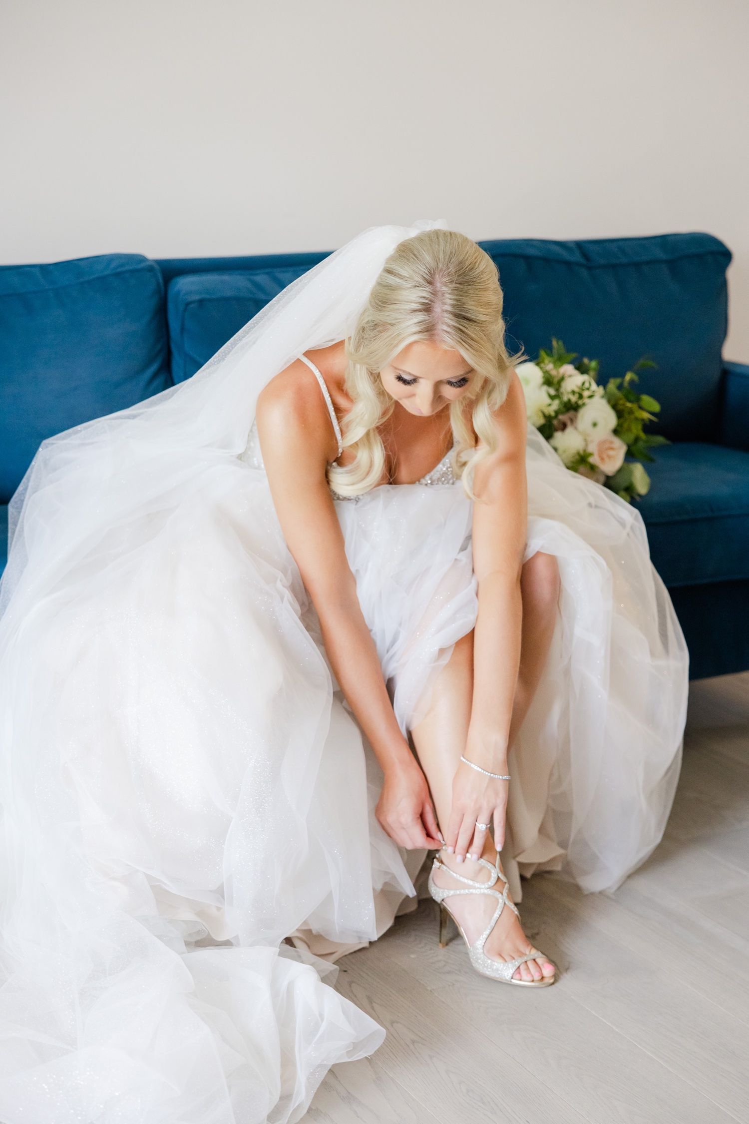 Close-up shot of bride in white gown adjusting her strappy wedding shoes on blue sofa.