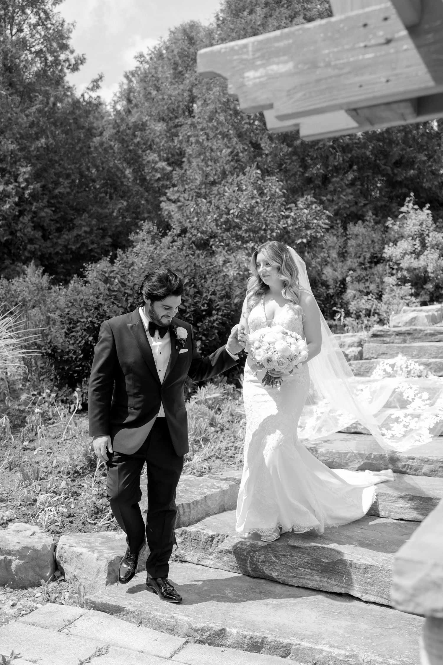 A black and white photo captures a couple walking down rustic stone steps in a garden setting.