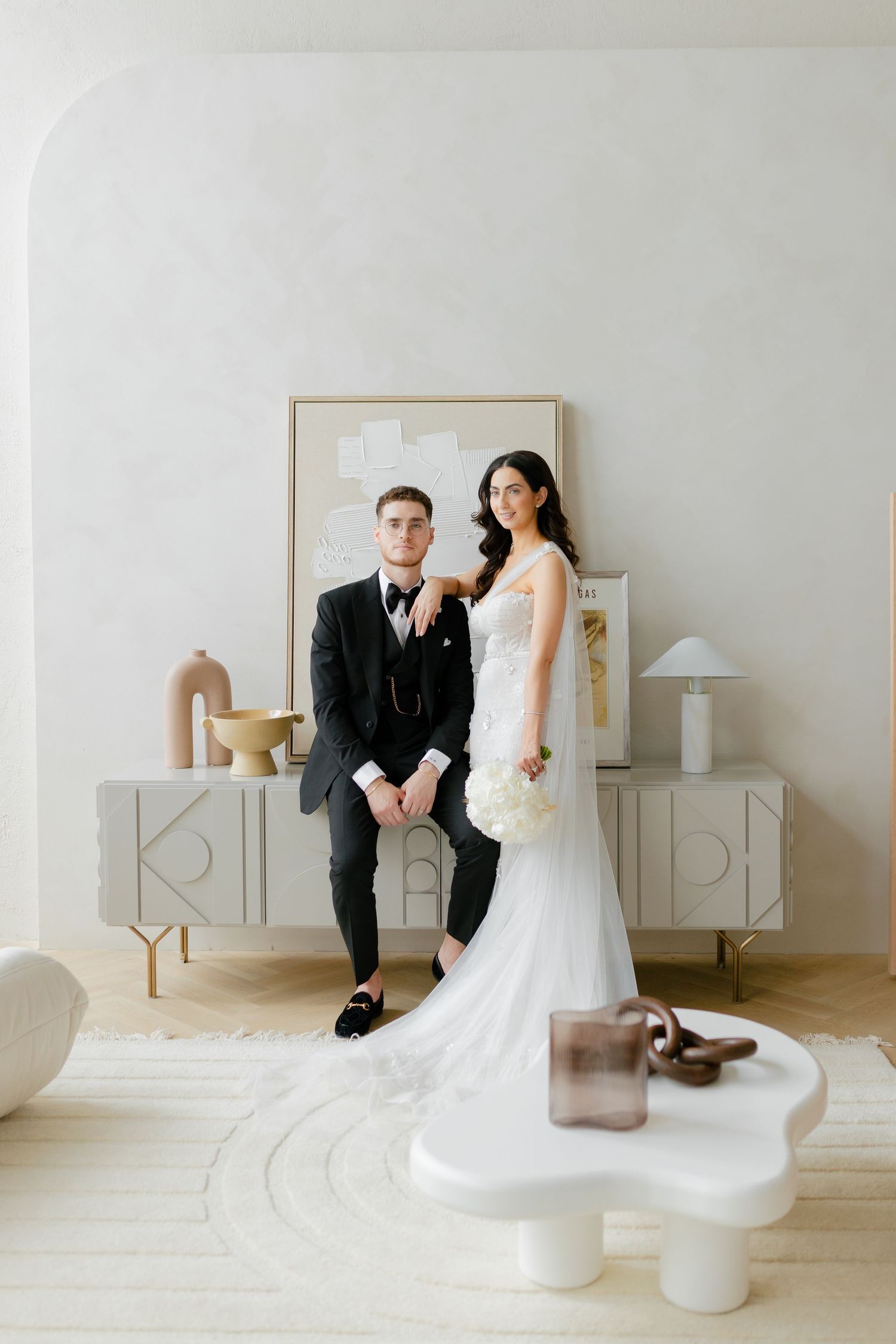 Modern wedding portrait with couple posed on white furniture against neutral minimalist decor.