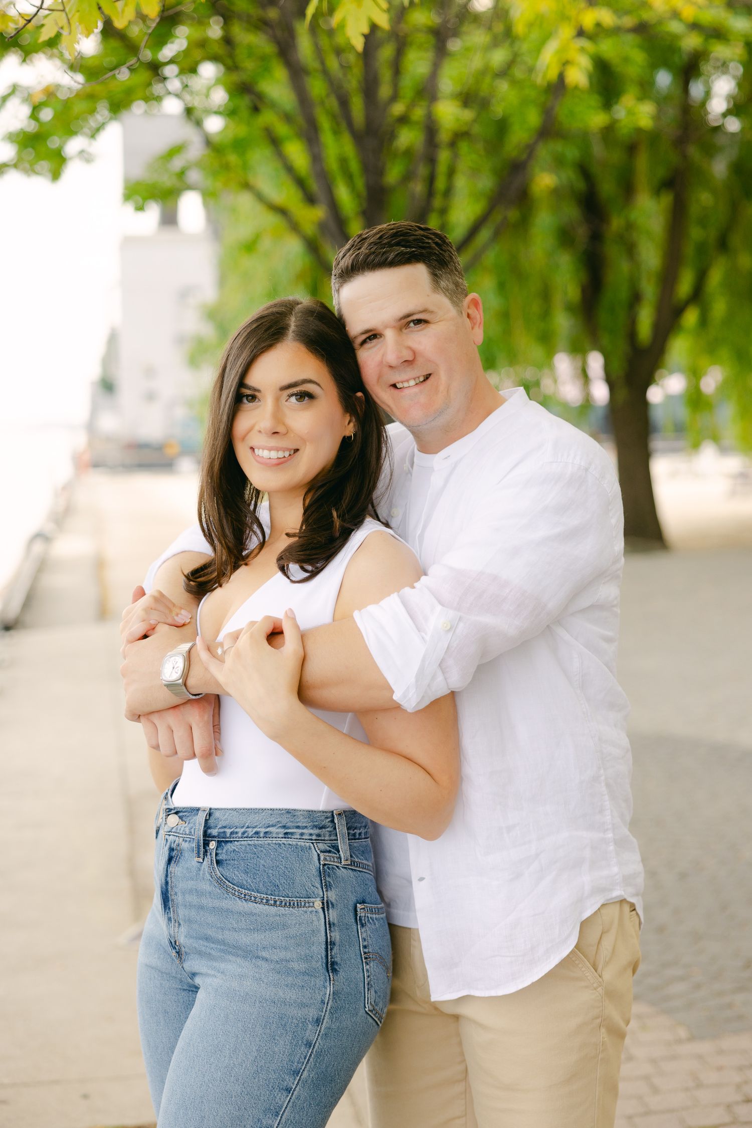 A couple embraces while standing outdoors near green trees, wearing casual white and denim outfits.