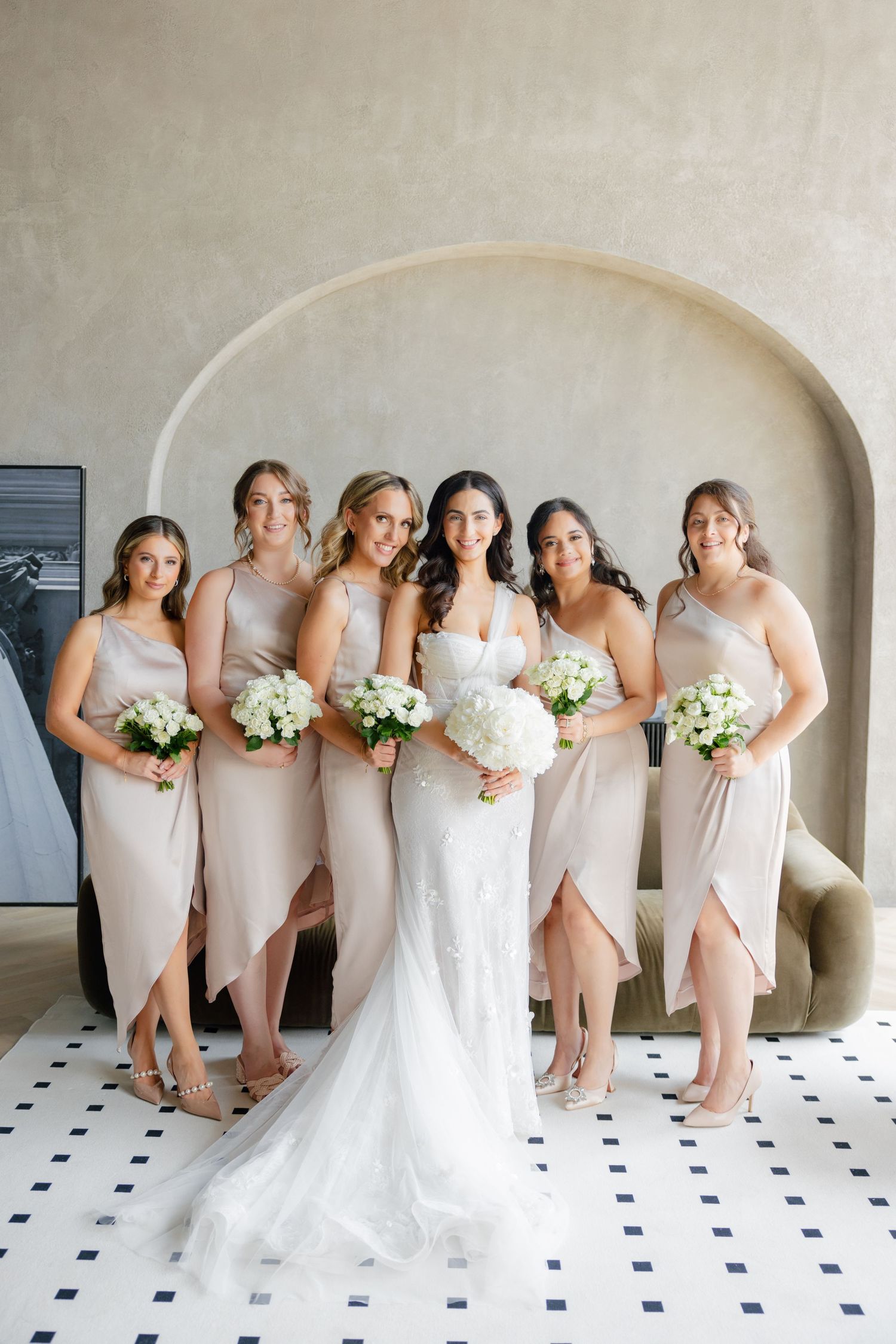 Bride stands centered among bridesmaids in blush dresses holding white floral bouquets.