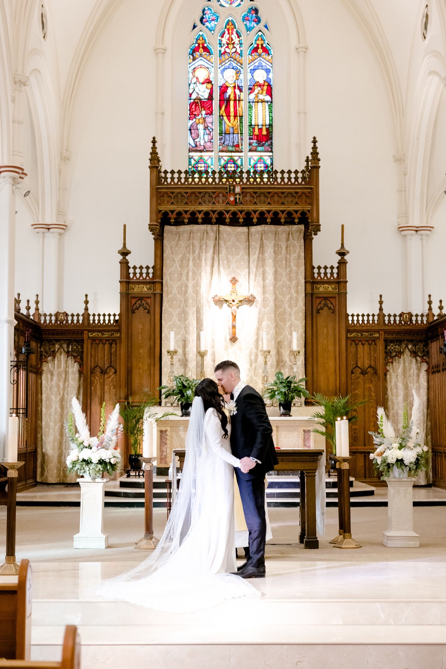 A wedding ceremony in a beautiful Gothic church with stained glass windows, ornate wooden altar screen and white floral arrangements.