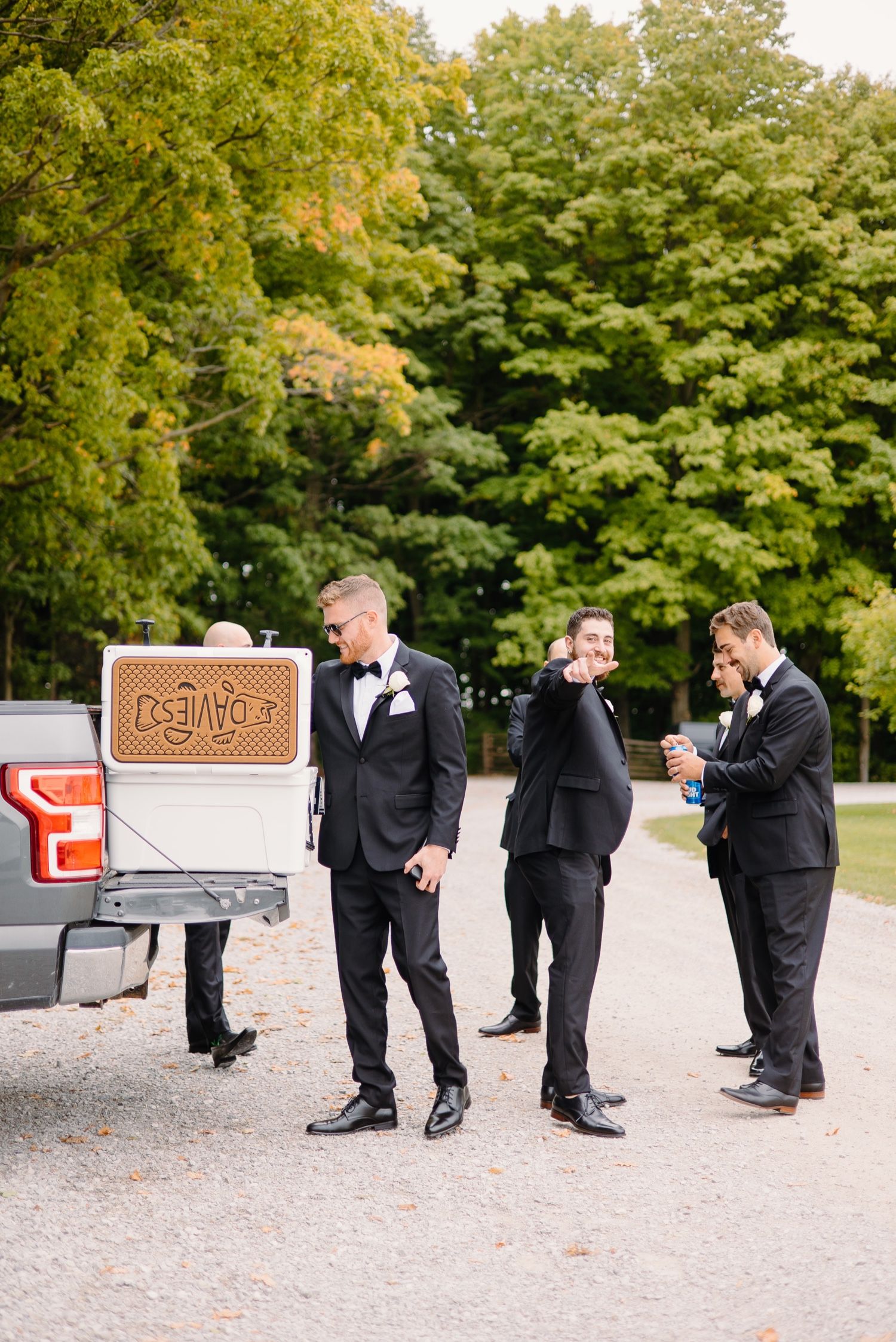 Groomsmen in tuxedos gather near pickup truck with cooler during wedding day.