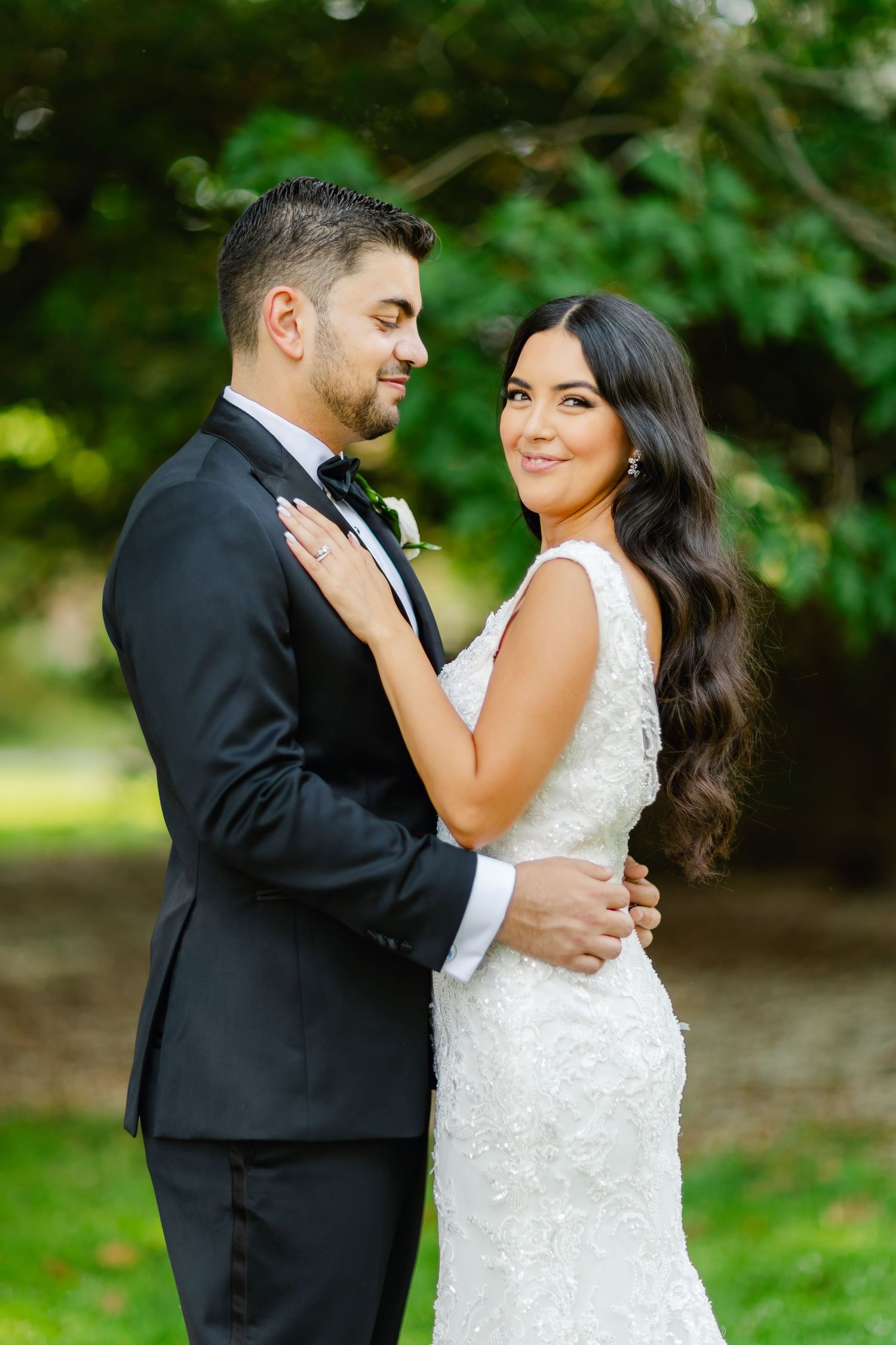 A romantic wedding couple embraces outdoors in a garden setting, with the bride in white lace and groom in black tuxedo.