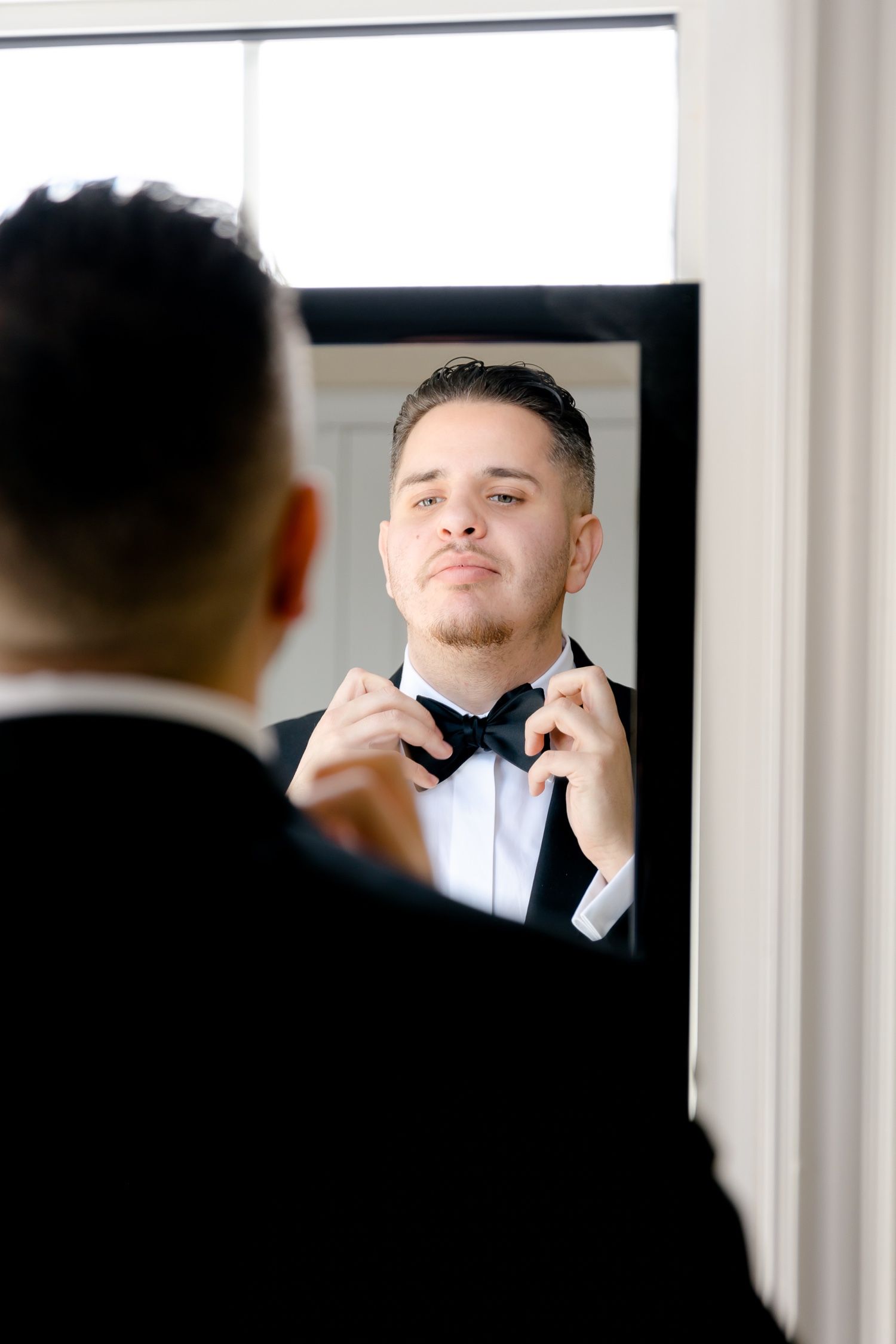 Groom adjusts bowtie while looking in mirror during wedding preparations.