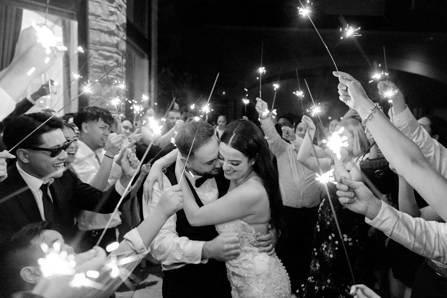 Black and white wedding photo showing couple embracing under sparkler arch at night.