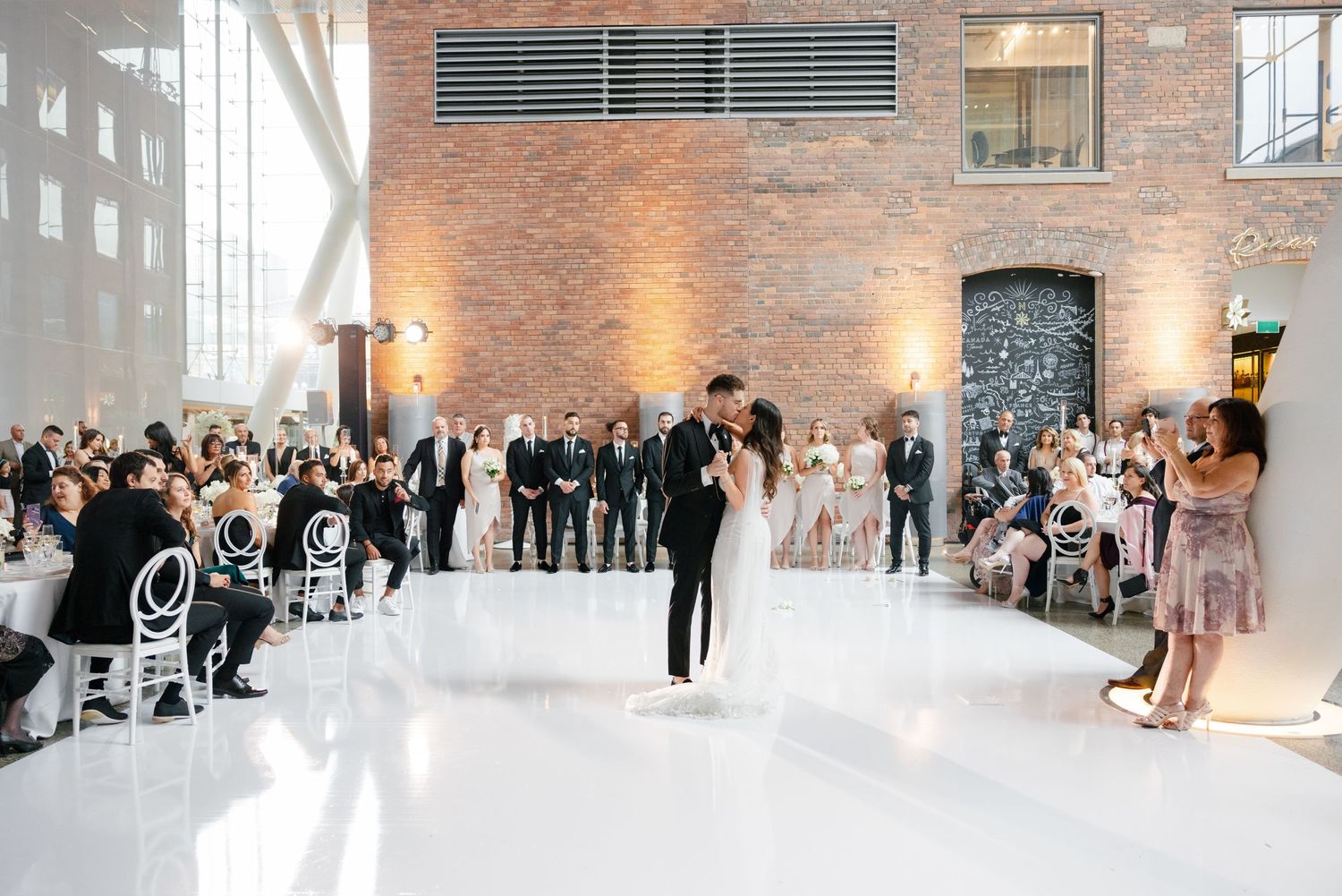 A newly married couple dancing in a bright industrial venue with exposed brick walls and wedding guests looking on.