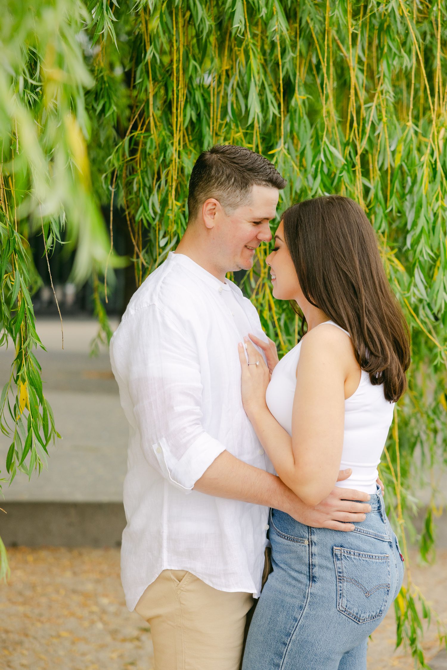 Loving couple embraces under cascading willow branches near water.