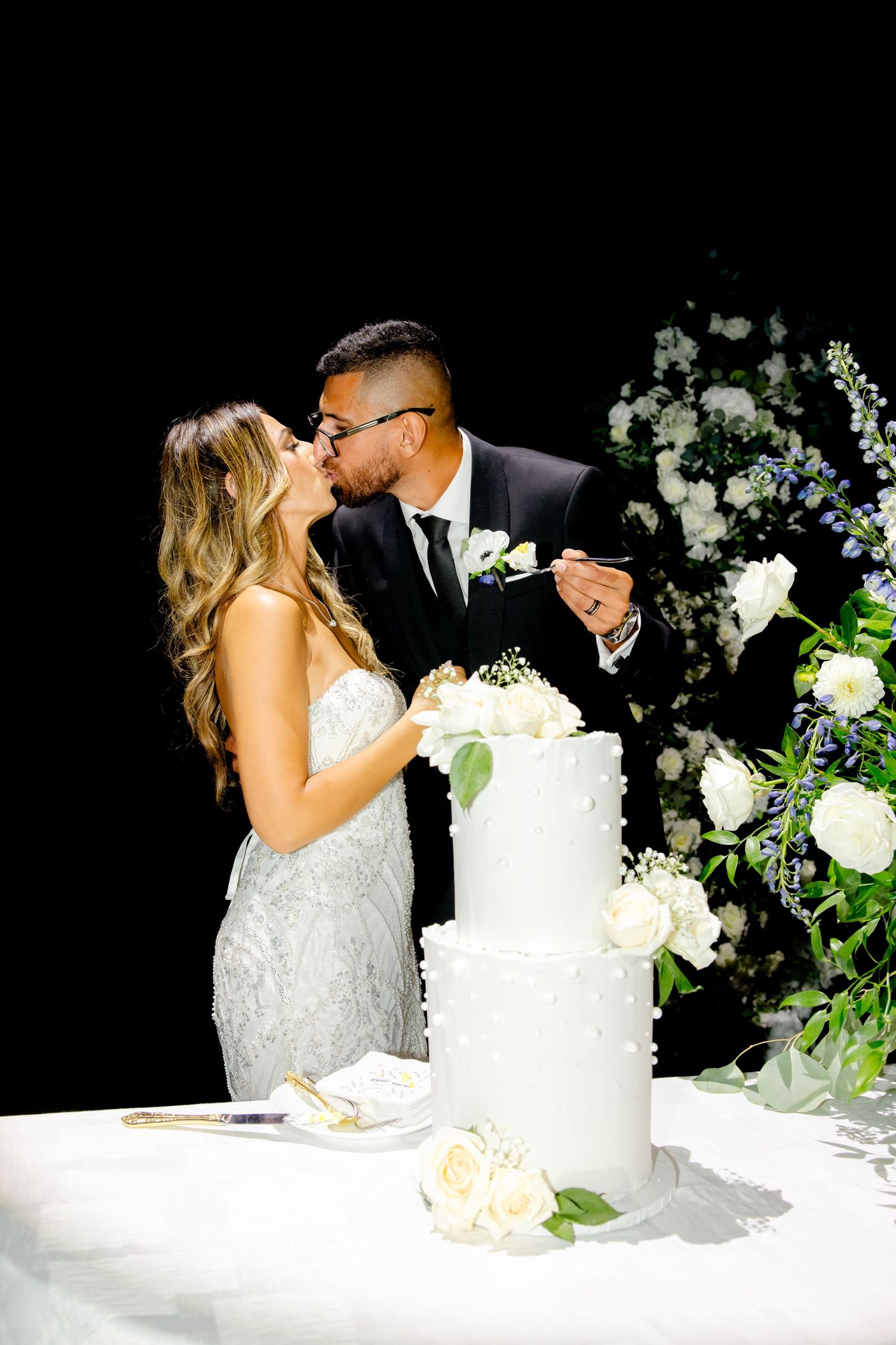 Romantic kiss shared during cake cutting with elegant white floral backdrop.