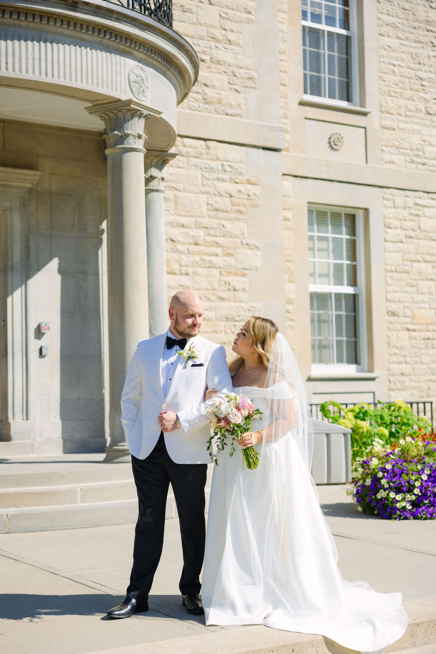 Sunlit wedding portrait in front of limestone mansion with classical columns and purple flowers.