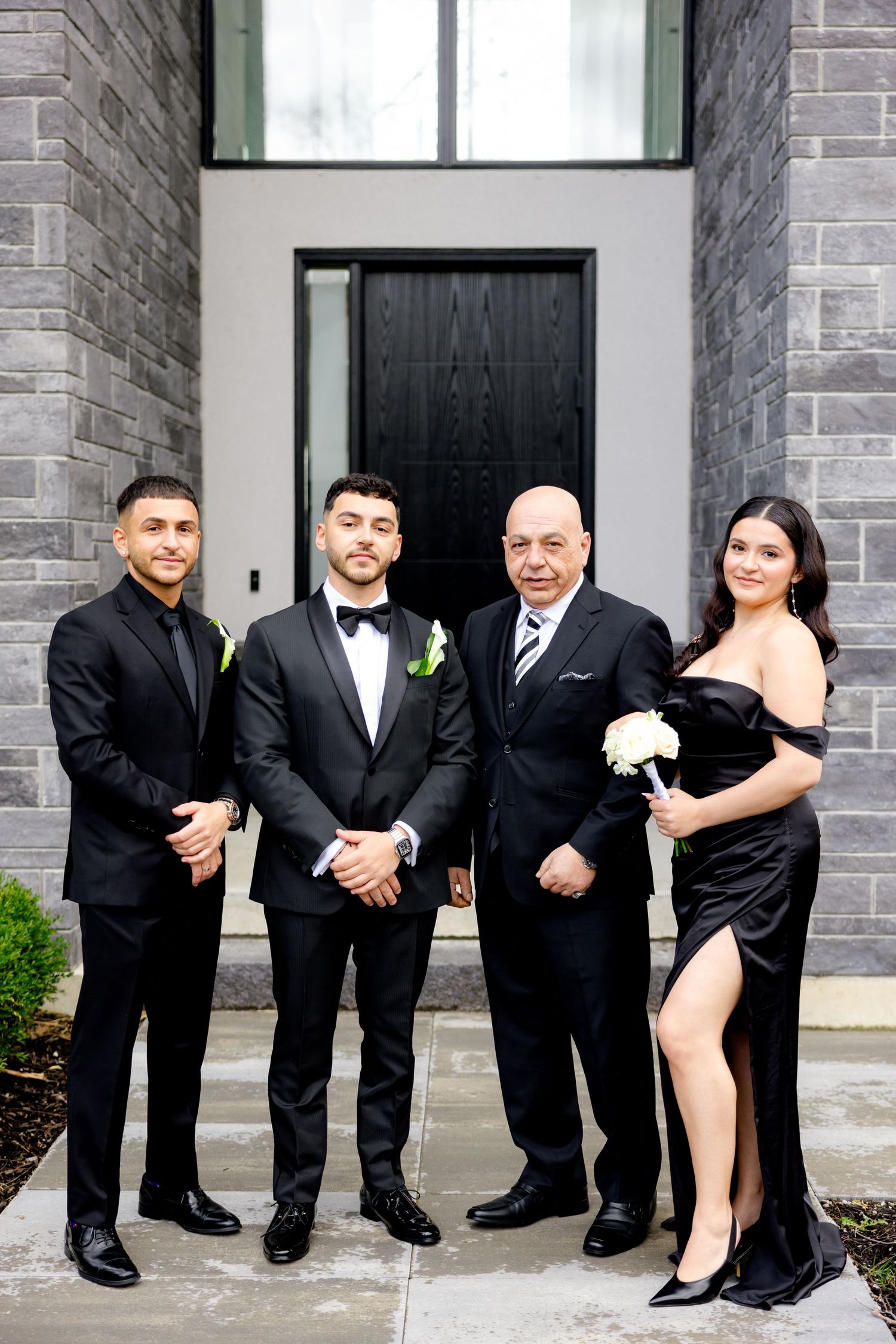 Wedding party in formal black attire poses in front of modern stone building entrance.